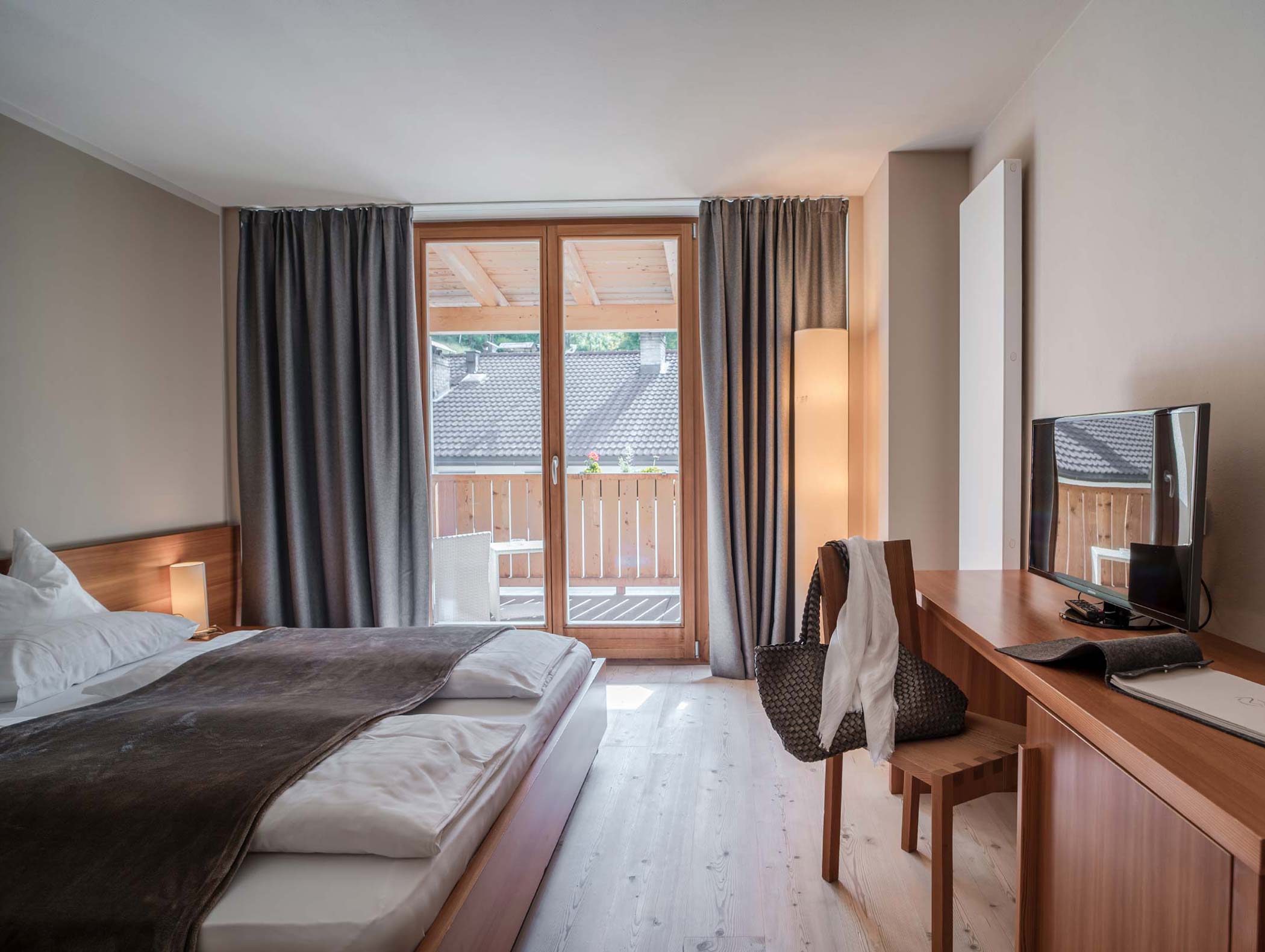 Moderna camera d'albergo con letto matrimoniale, scrivania, TV e balcone con ringhiera in legno e posti a sedere all'aperto. - Hotel Moosmair