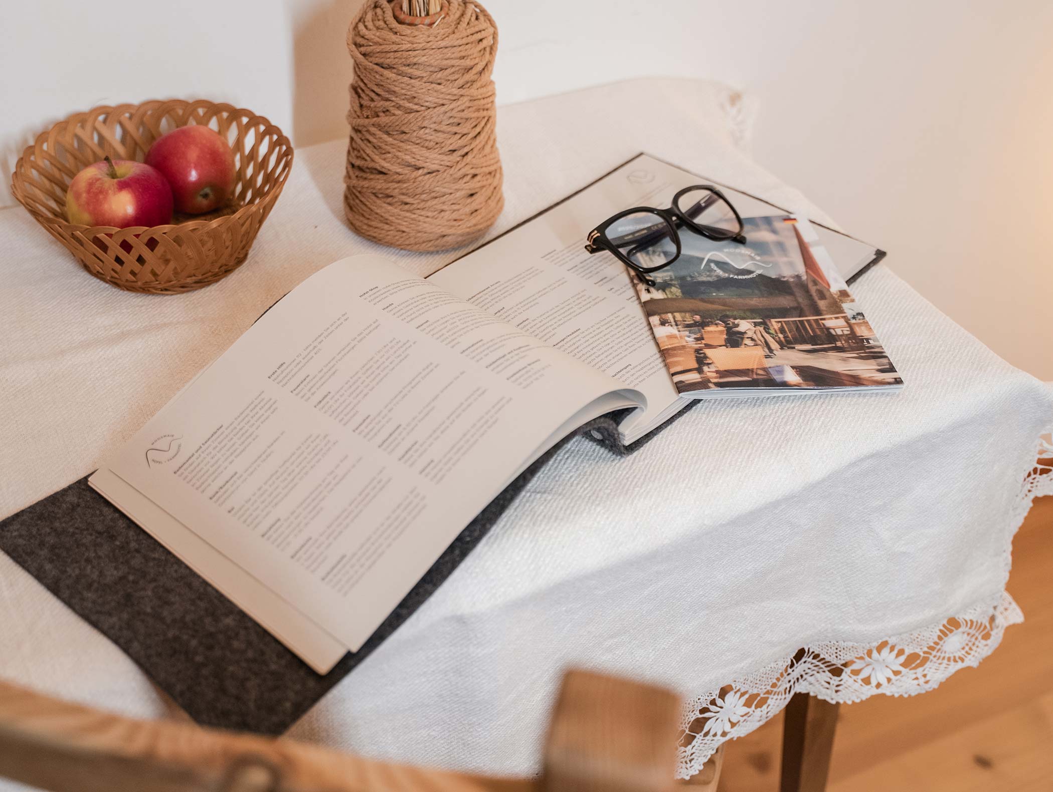 Libro aperto, bicchieri e mele su un tavolo rivestito di pizzo con un rocchetto di spago in un ambiente accogliente. - Hotel Moosmair