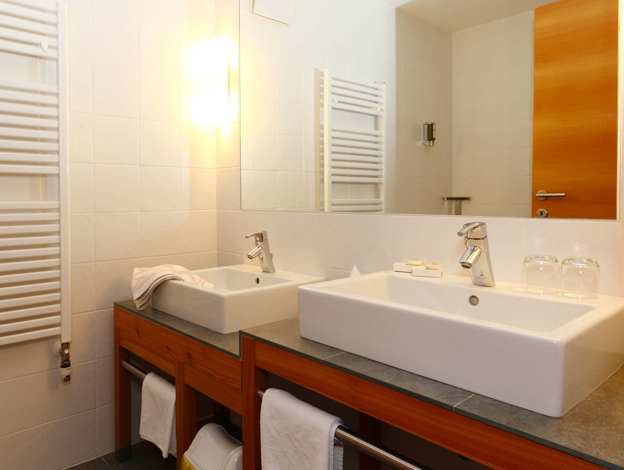 Bagno moderno con due lavandini quadrati, un grande specchio, asciugamani e un'illuminazione intensa. - Hotel Moosmair