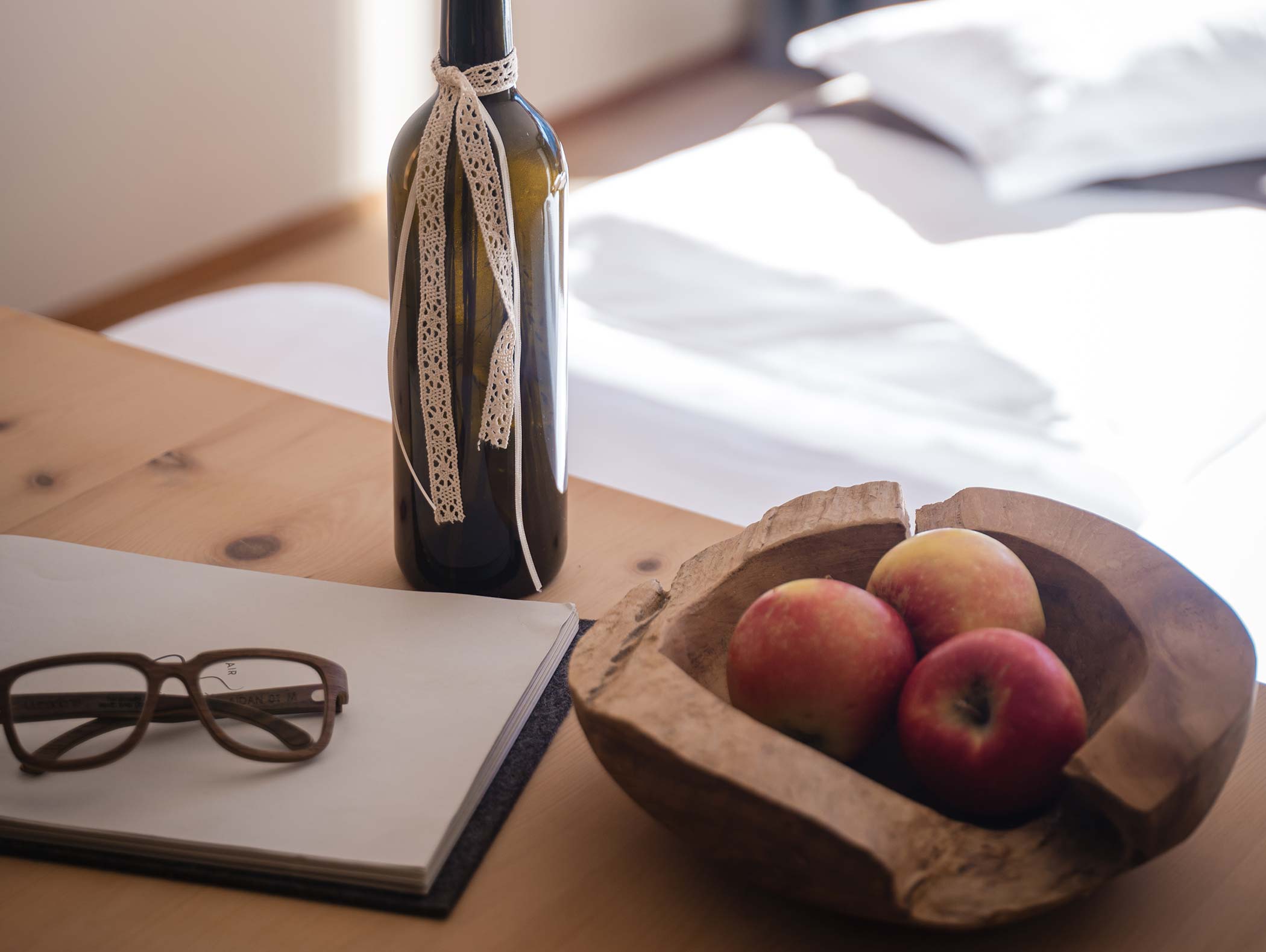 Una ciotola di legno con mele, una bottiglia di vino con un nastro di pizzo, bicchieri e un quaderno su un tavolo di legno. - Hotel Moosmair