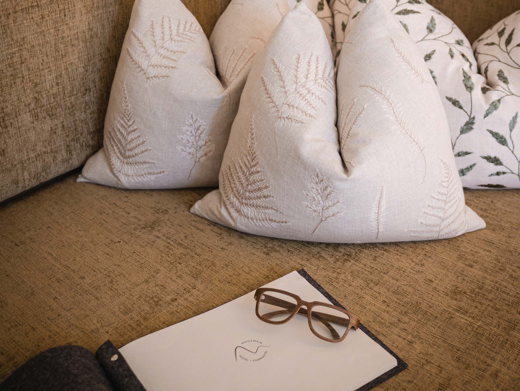 Divano beige con cuscini a forma di foglia, occhiali marroni e un blocco note sulla seduta. - Hotel Moosmair
