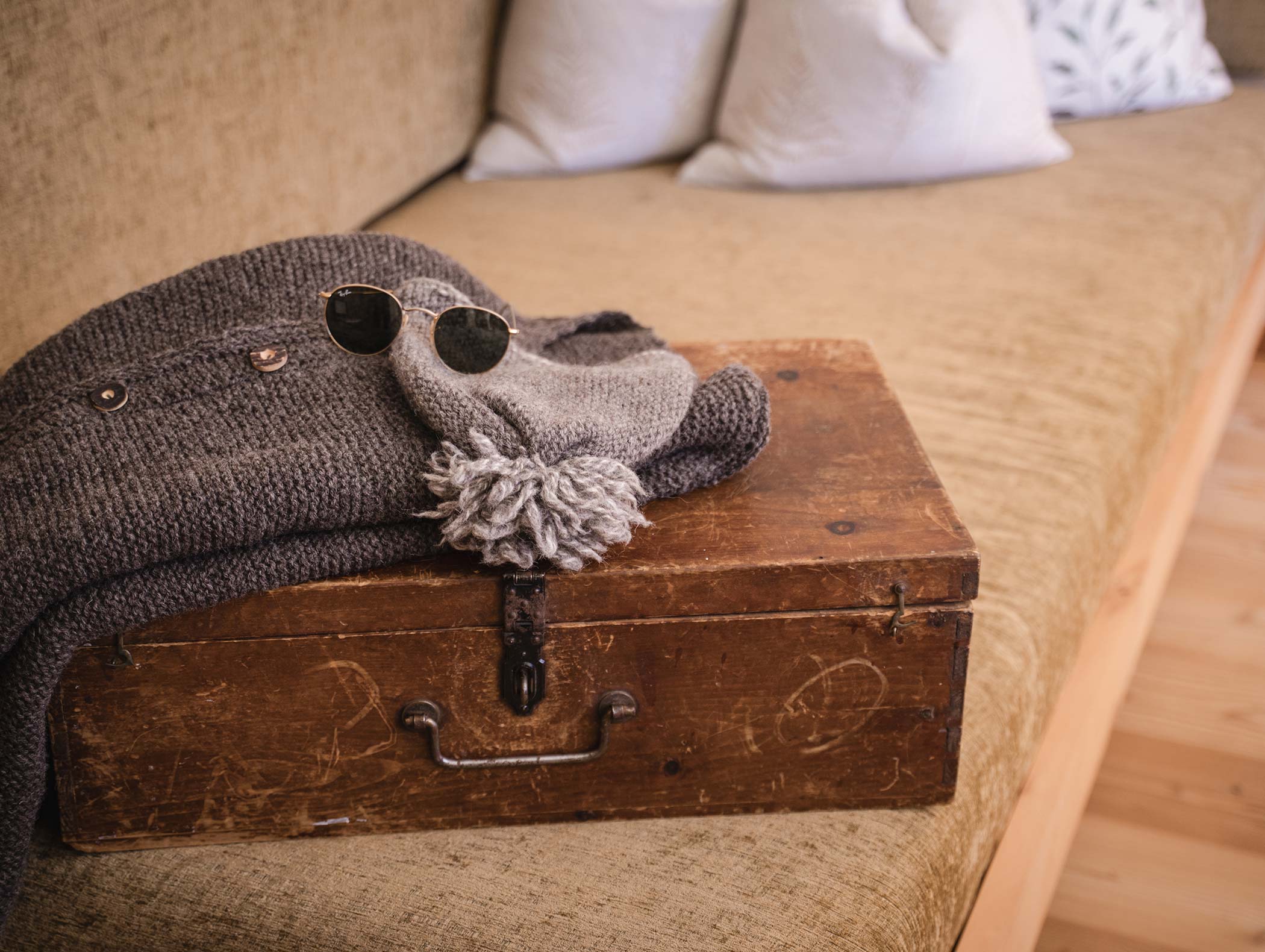 Una valigia di legno con maglione grigio, cappello a maglia e occhiali da sole su un divano beige. - Hotel Moosmair