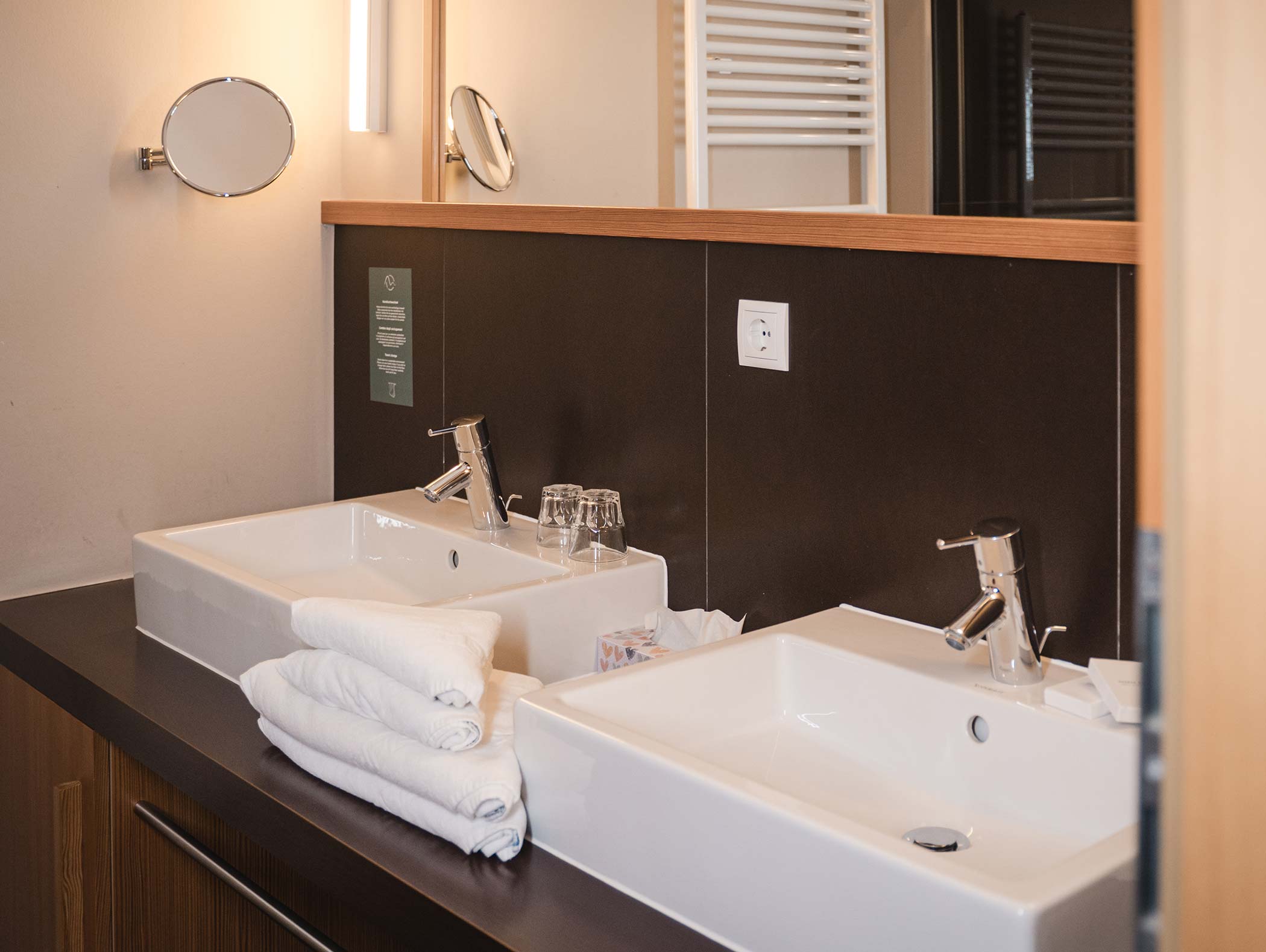 Due lavandini bianchi con rubinetti cromati, asciugamani, bicchieri e specchi impilati su un piano di lavoro scuro. - Hotel Moosmair