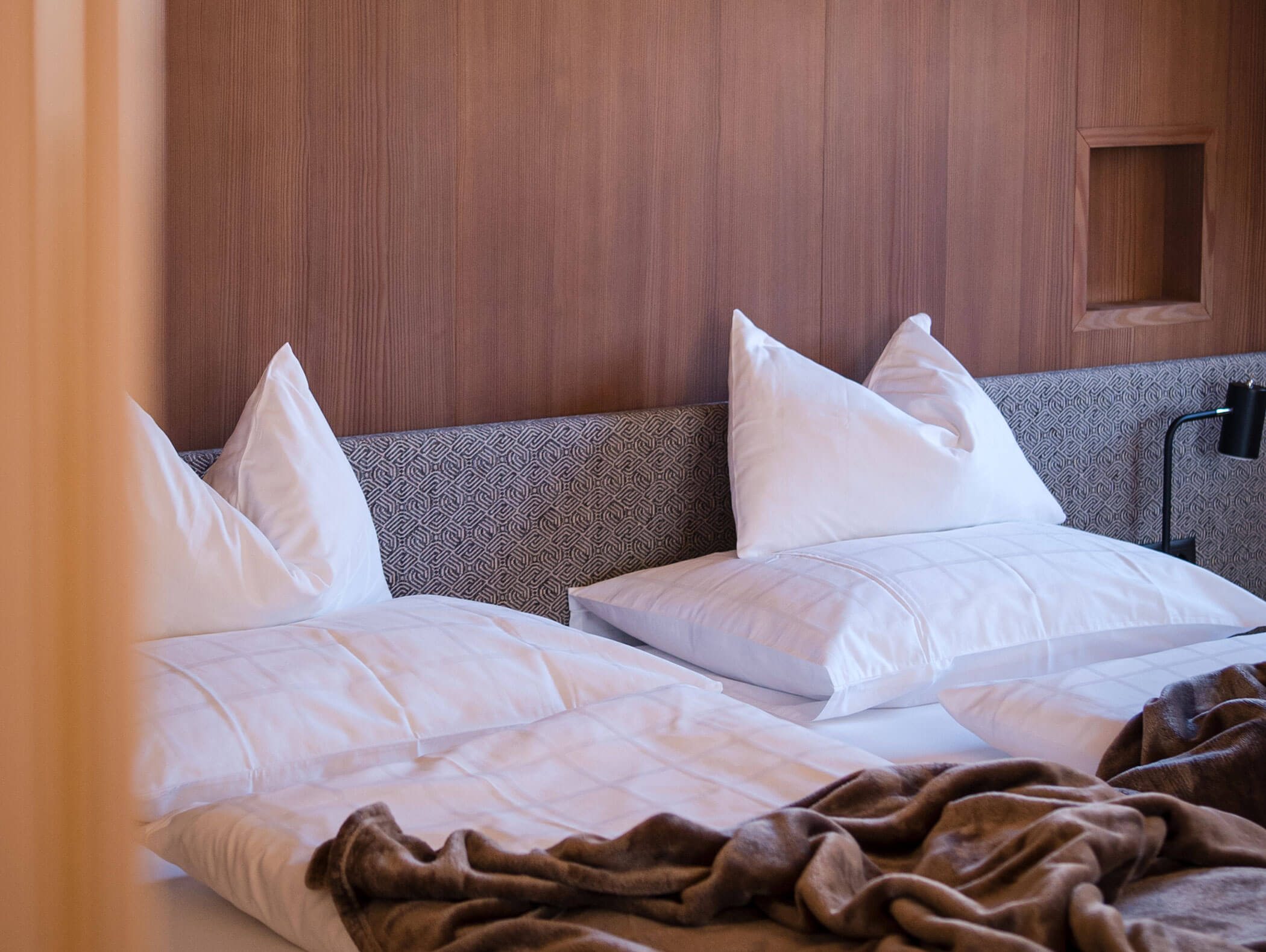 Due letti con cuscini e piumoni bianchi, una morbida coperta marrone e una testiera in legno in una stanza accogliente. - Hotel Moosmair