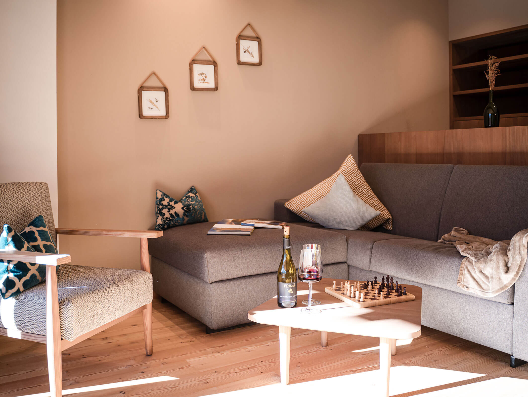 Accogliente soggiorno con divano grigio, poltrona, vino, set di scacchi e opere d'arte su una parete beige. - Hotel Moosmair