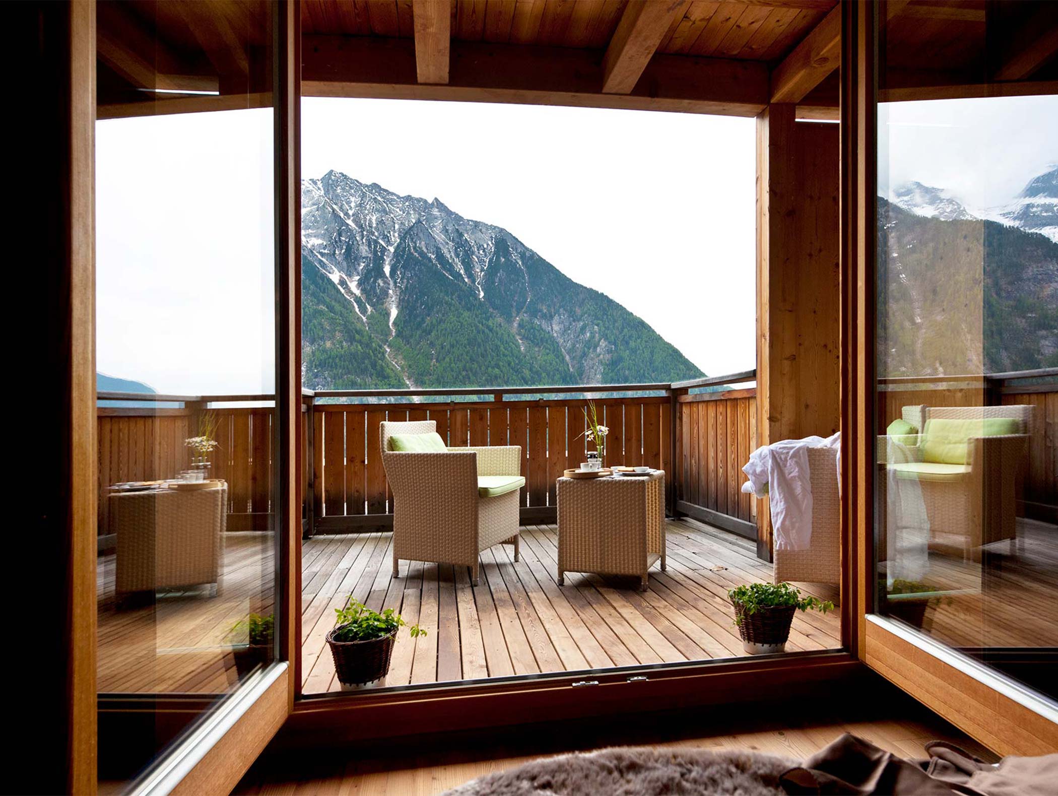 La terrazza in legno con sedie e tavolini si affaccia su un paesaggio montano, visibile attraverso grandi porte di vetro. - Hotel Moosmair