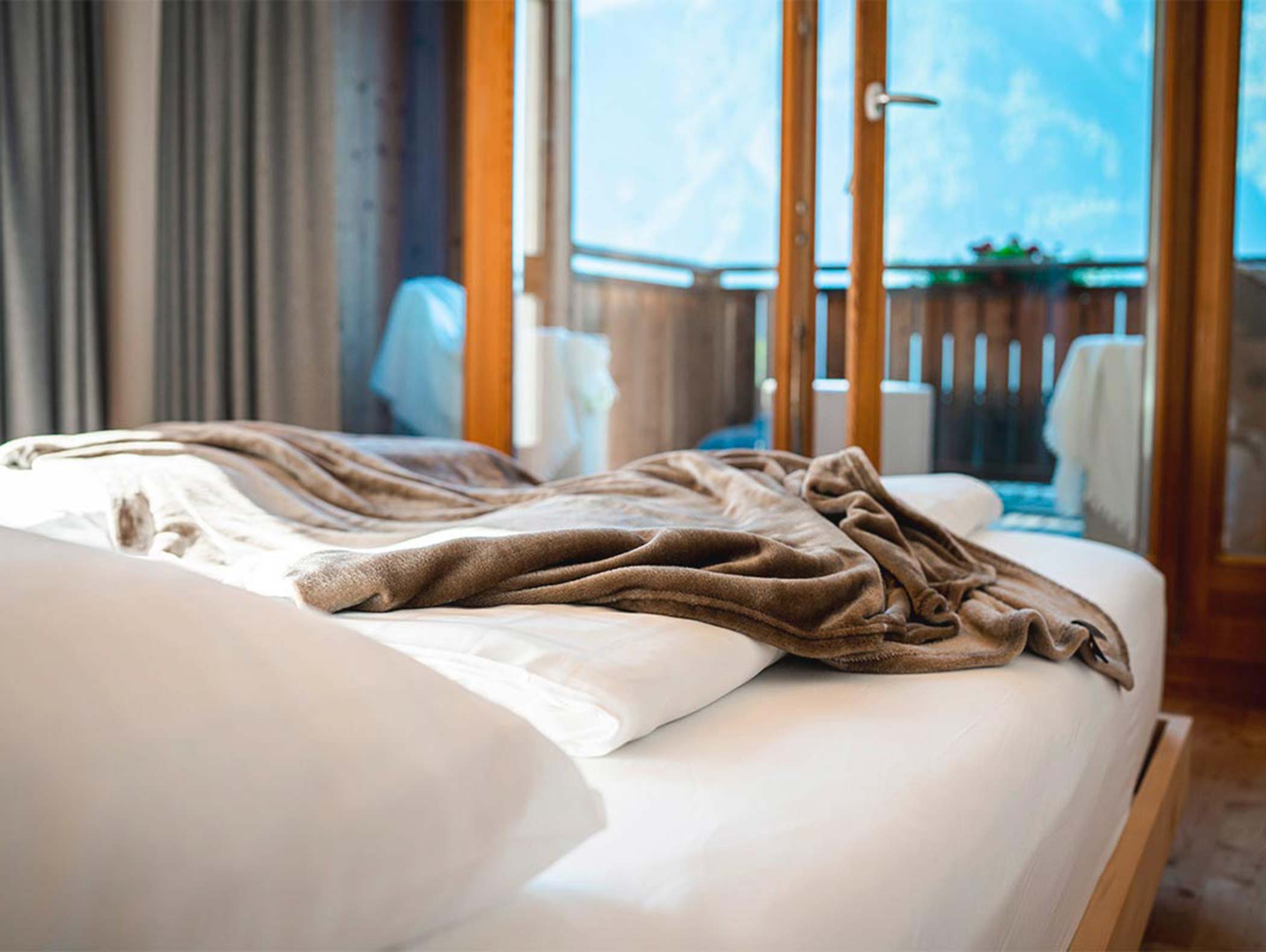 Un letto accogliente con lenzuola bianche e una coperta marrone, vicino a porte aperte che conducono a un balcone soleggiato. - Hotel Moosmair