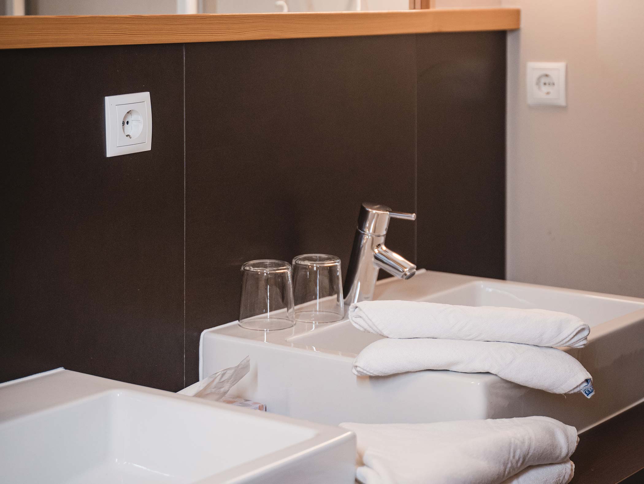 Un lavandino da bagno moderno con due bicchieri, asciugamani bianchi e un rubinetto cromato su uno sfondo scuro. - Hotel Moosmair
