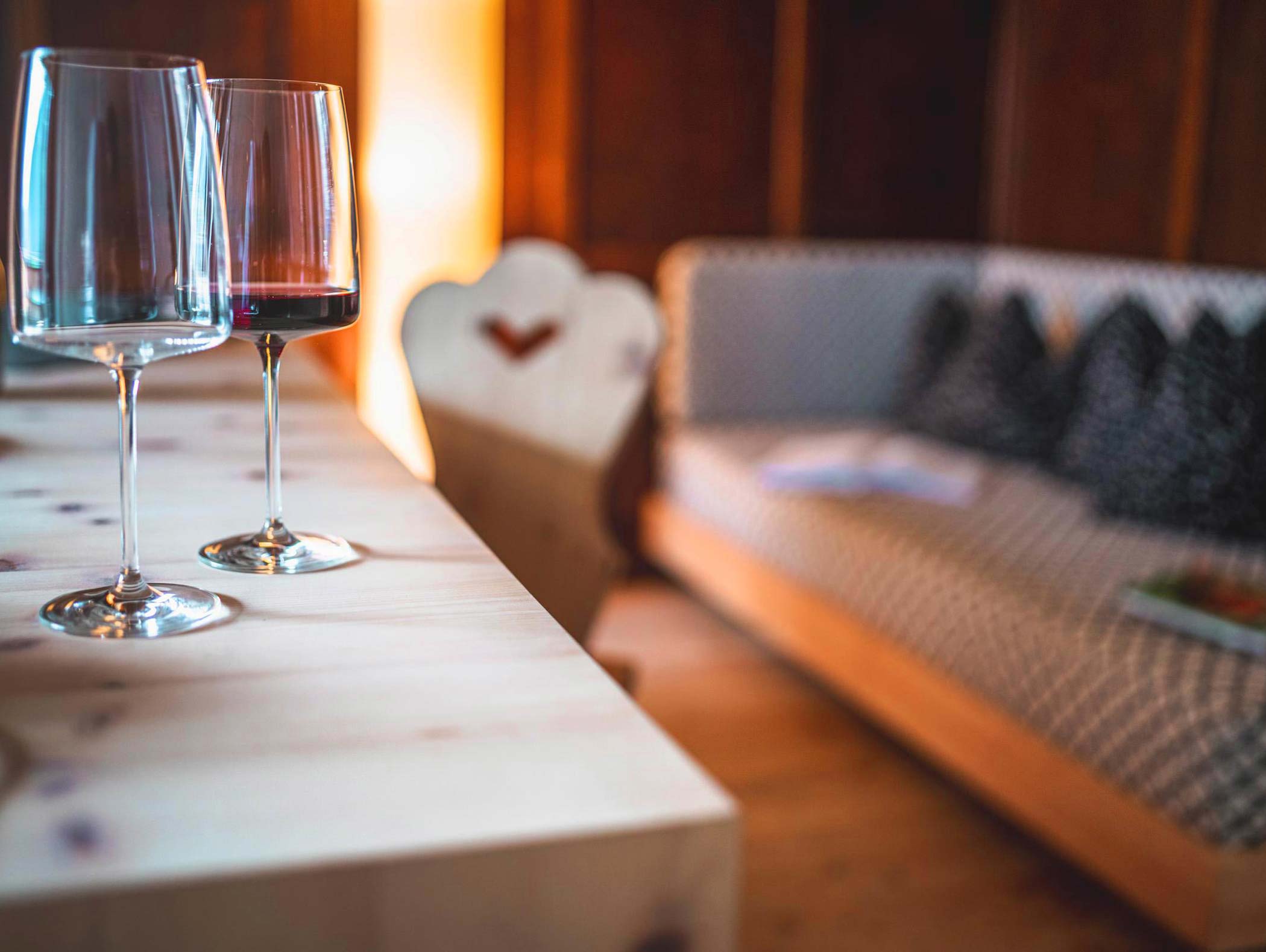 Due bicchieri da vino, uno con vino rosso, su un tavolo di legno accanto a un divano accogliente in una stanza dalla luce calda. - Hotel Moosmair