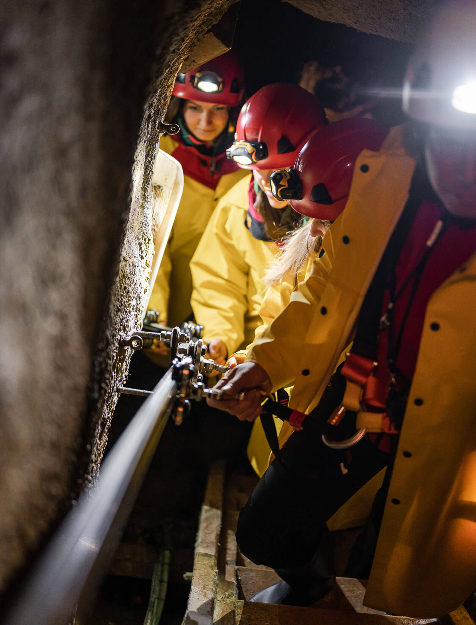 Tre persone con giacche gialle e caschi rossi lavorano insieme all'attrezzatura in un tunnel stretto e roccioso. - Hotel Moosmair