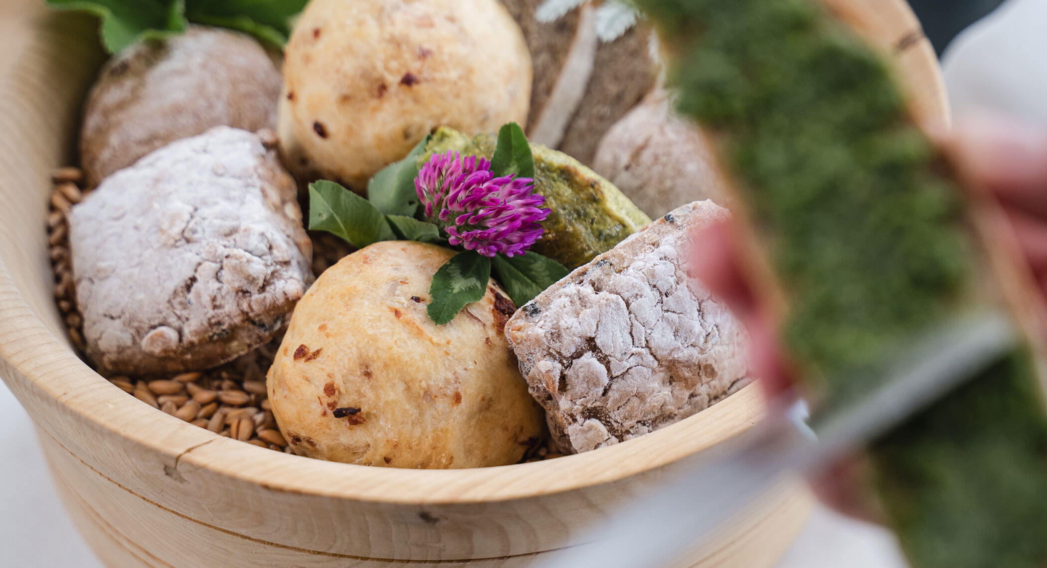 Verschiedene handwerkliche Brötchen in einer Holzschale, garniert mit einer lila Blume und grünen Blättern. - Hotel Moosmair