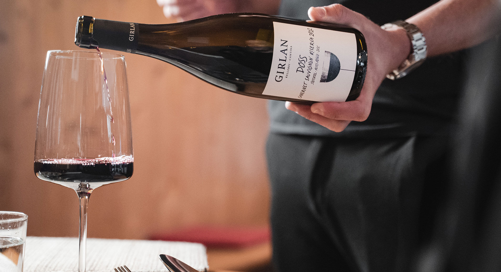 Eine Person schenkt Rotwein aus einer Flasche mit der Aufschrift Girlan in ein Glas auf einem Esstisch ein. - Hotel Moosmair