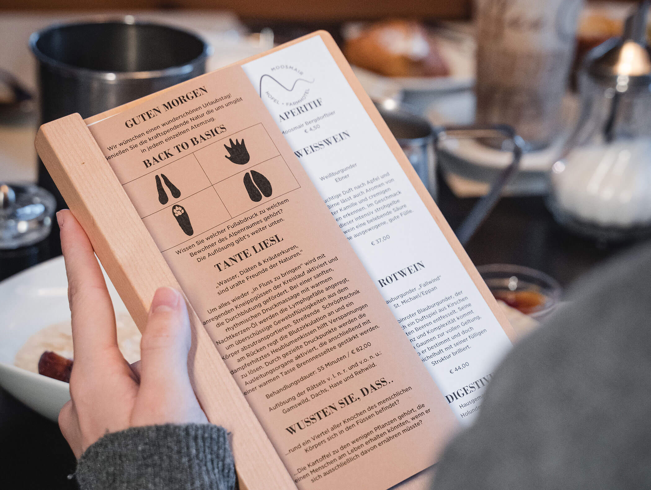 Person, die an einem gedeckten Tisch eine Frühstückskarte in deutscher Sprache mit Gerichten und Illustrationen in der Hand hält. - Hotel Moosmair