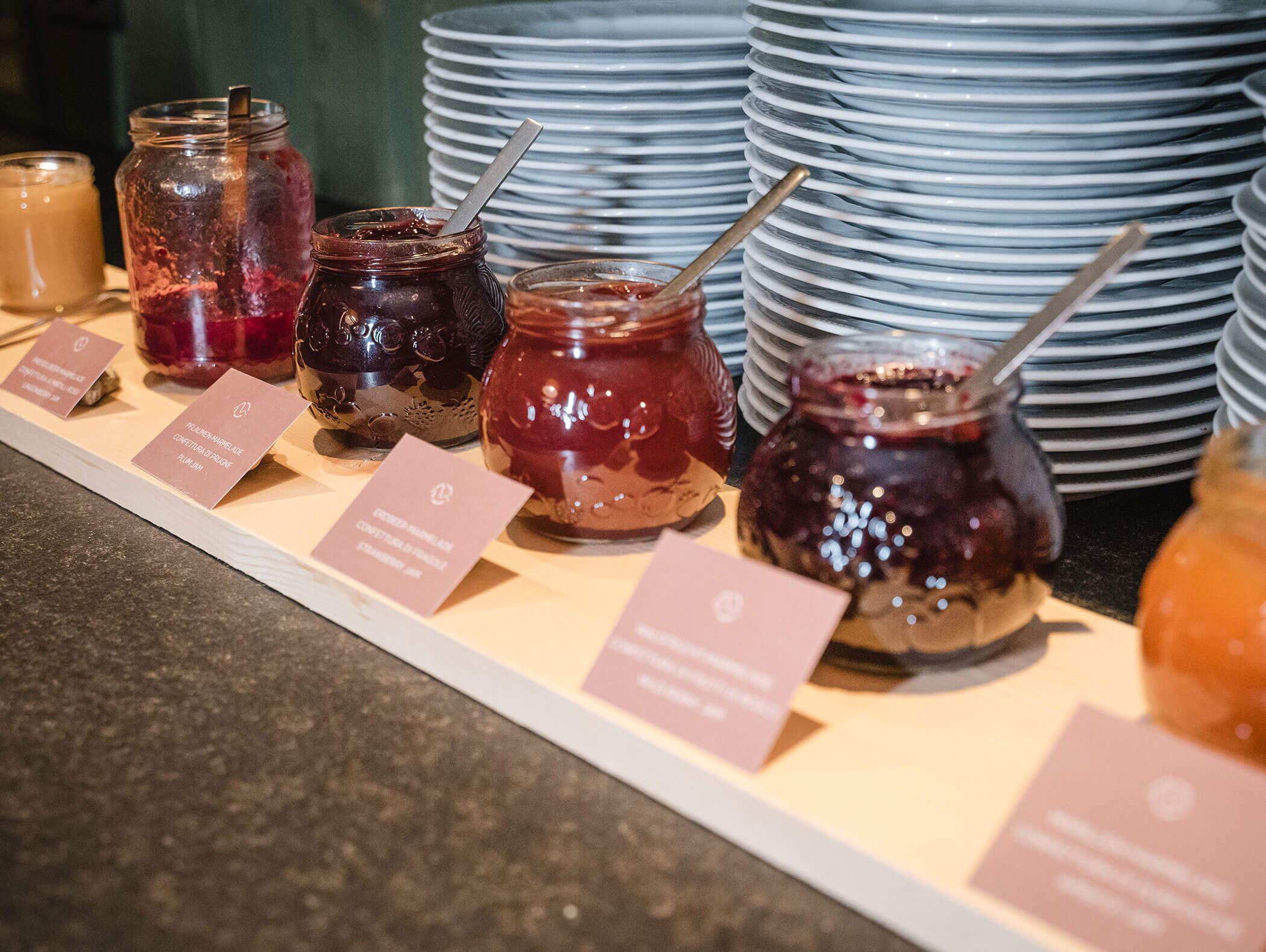 Fünf Gläser mit verschiedenen Marmeladen und Löffeln, die auf einem Buffet vor gestapelten Tellern und beschrifteten Karten ausgestellt sind. - Hotel Moosmair