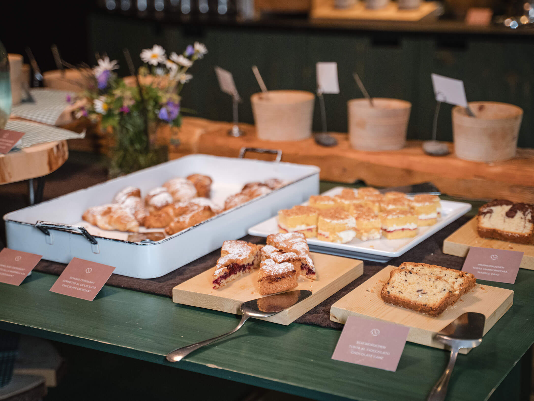 Gemischtes Gebäck und Kuchen auf Tabletts mit Servierutensilien, die auf einem Tisch neben einer Blumenvase ausgestellt sind. - Hotel Moosmair