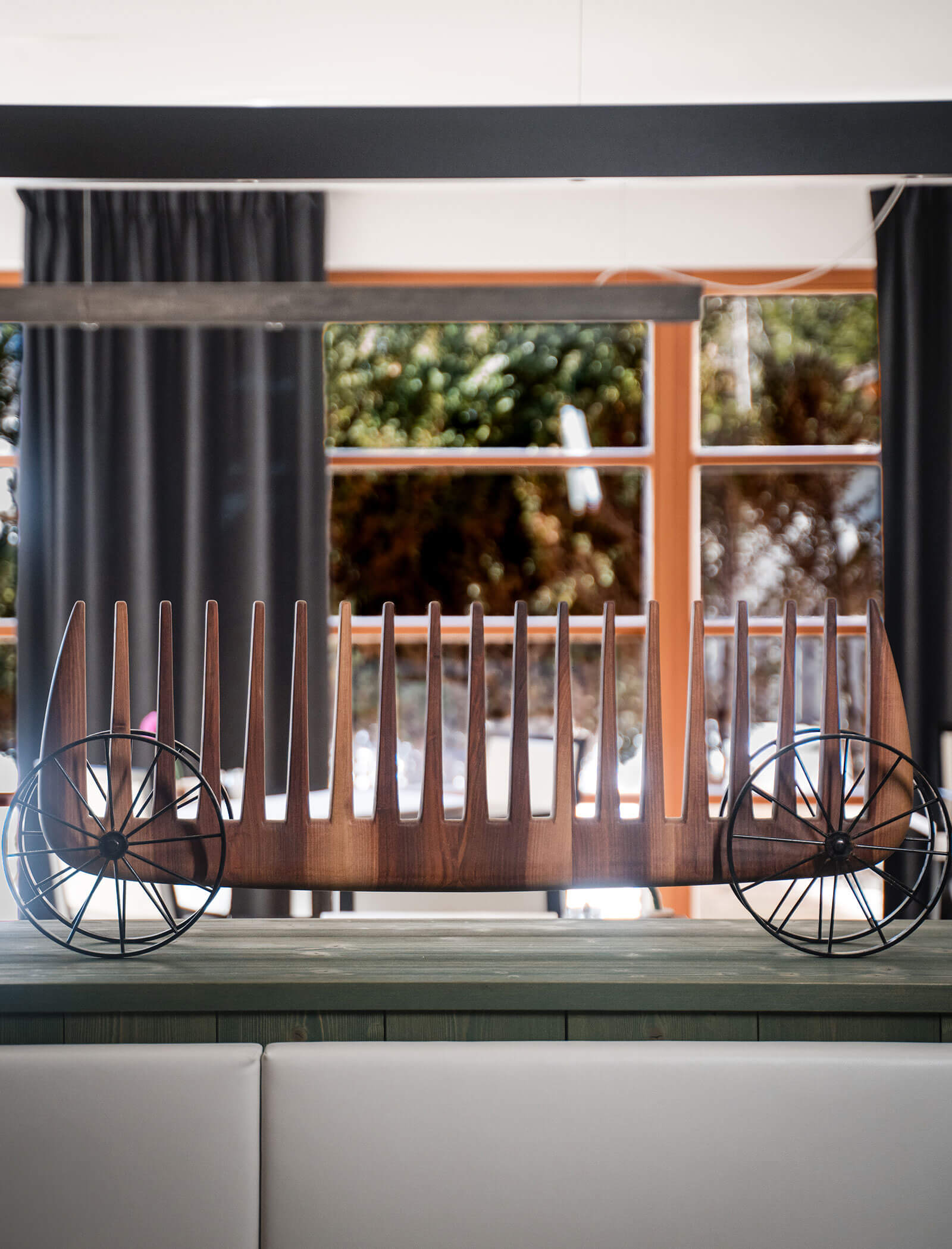 A modern wooden decorative piece with vertical slats and metal wheels, displayed indoors near large windows. - Hotel Moosmair