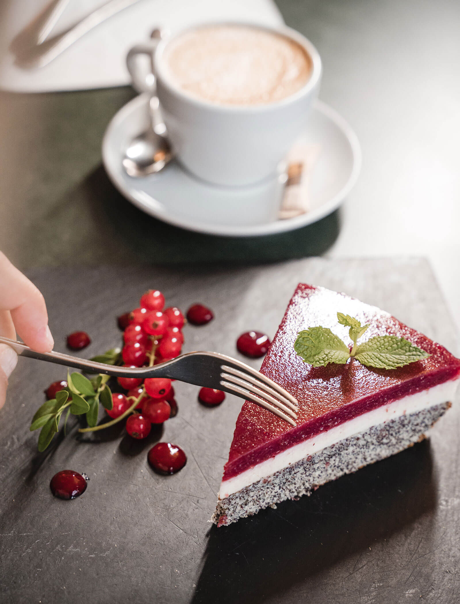 A fork cuts into a slice of berry mousse cake with mint, near red currants and a cup of coffee. - Hotel Moosmair