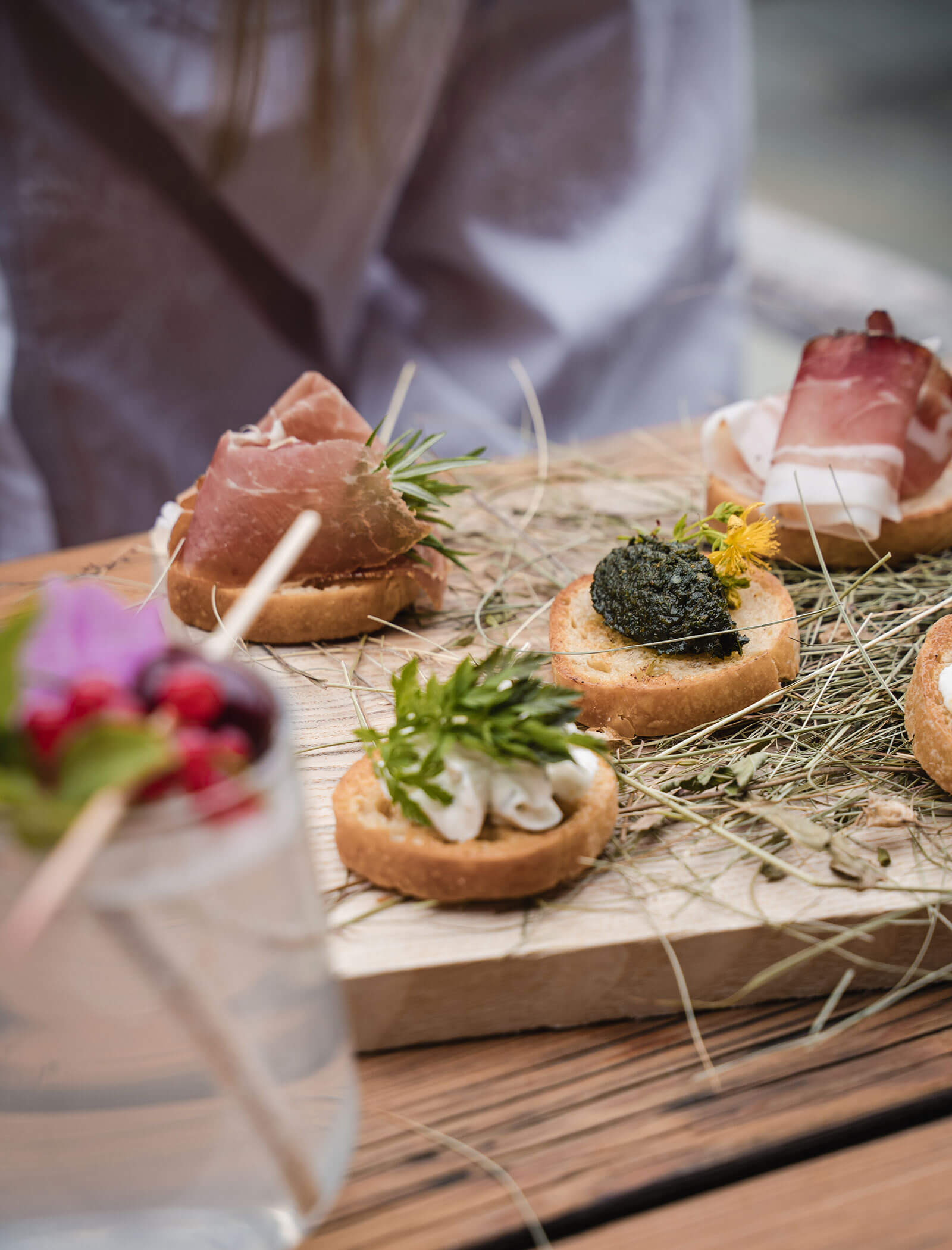 A wooden board with assorted gourmet canapés and herbs, next to a jar with a floral drink, on a rustic table. - Hotel Moosmair