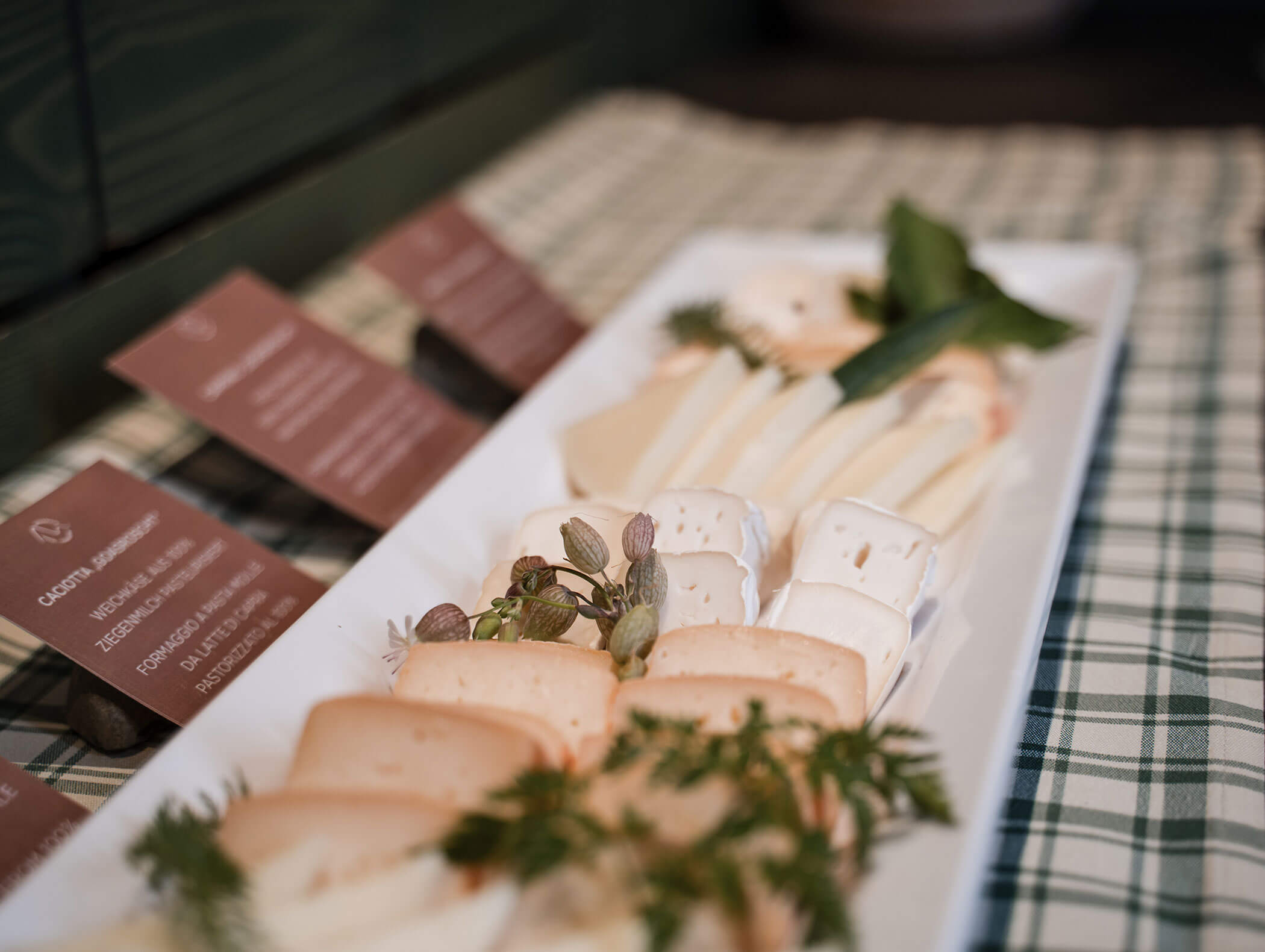A rectangular plate with assorted sliced cheeses and herbs on a checkered tablecloth. - Hotel Moosmair
