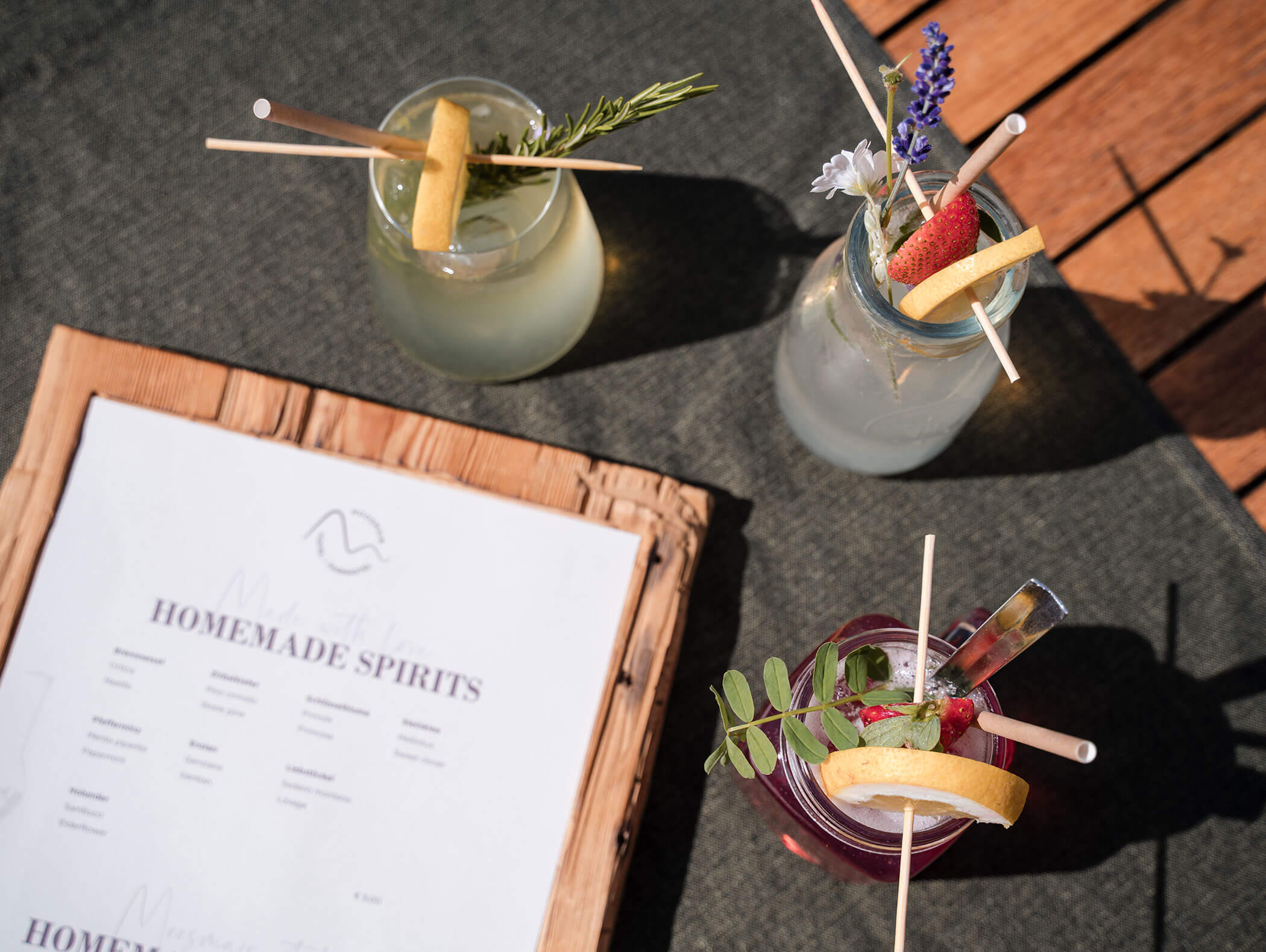 Three colorful cocktails with fruit and herb garnishes beside a homemade spirits menu on a woven placemat. - Hotel Moosmair
