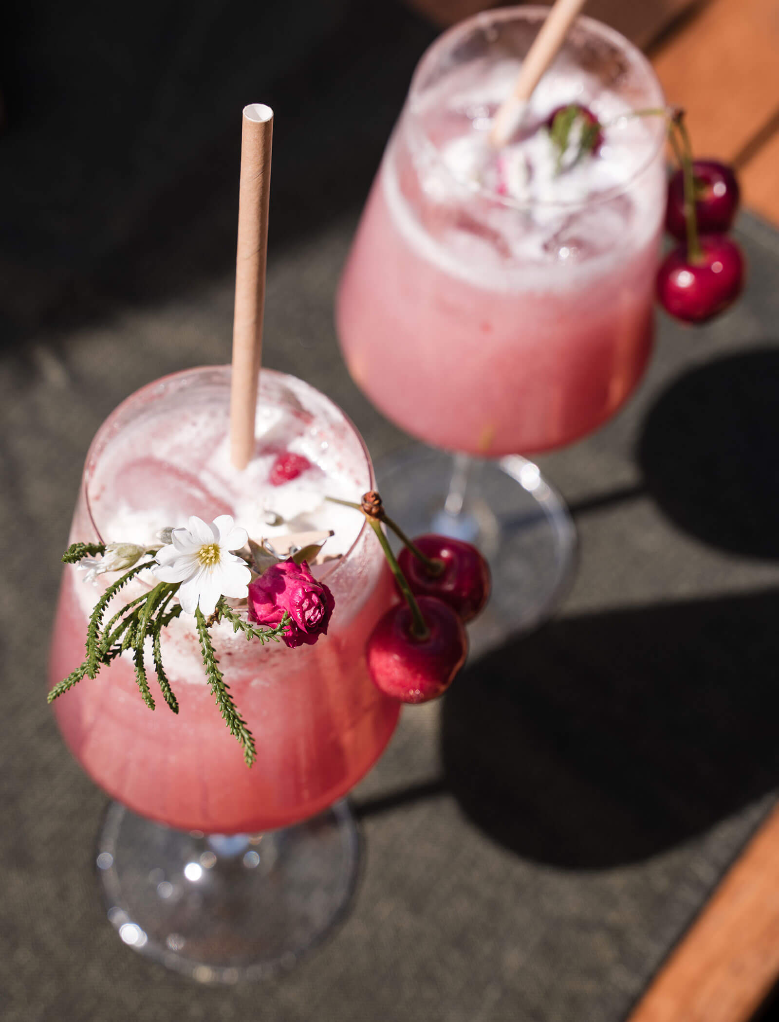 Two pink cocktails with ice, garnished with cherries and flowers, served in stemmed glasses with straws. - Hotel Moosmair