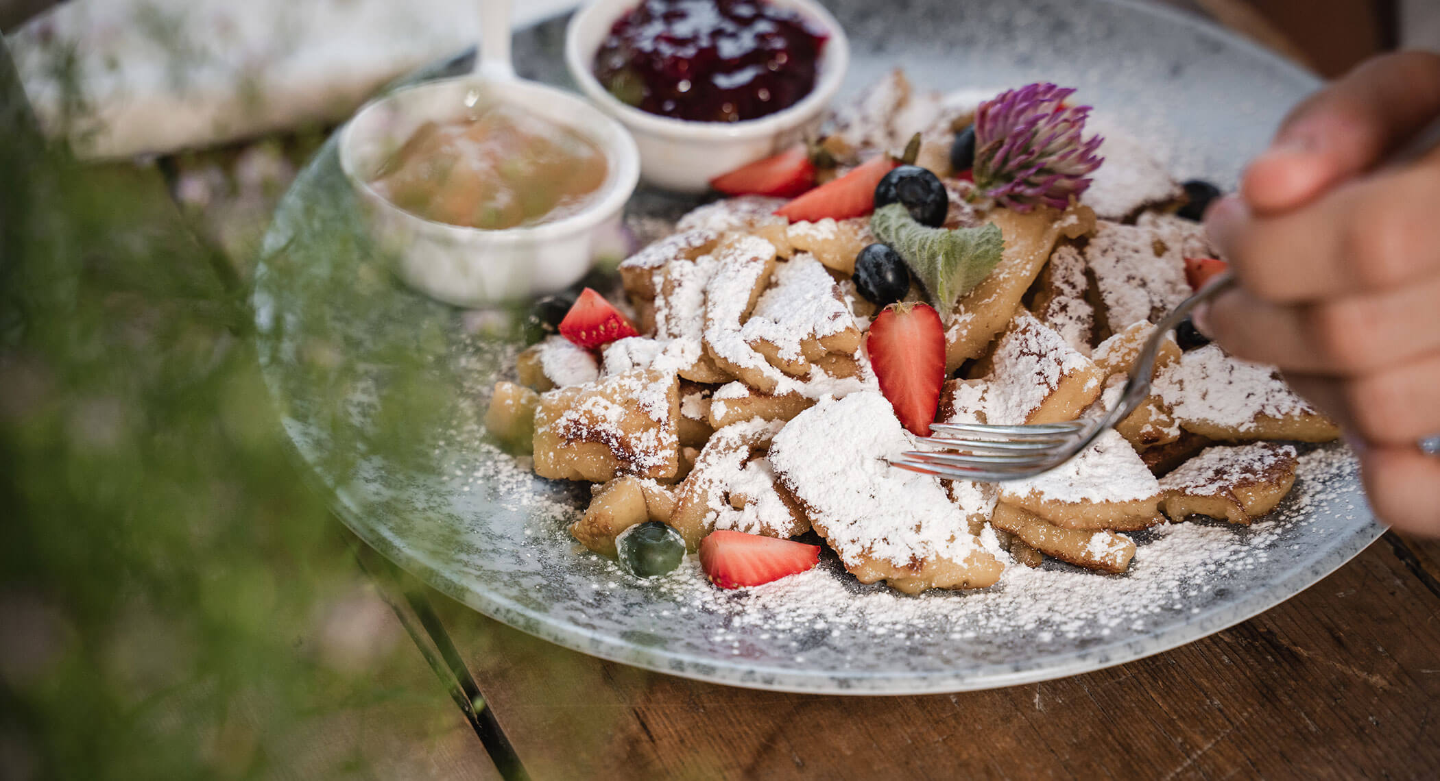 A plate of pancakes topped with powdered sugar, strawberries, blueberries, and flowers, with sauces on the side. - Hotel Moosmair