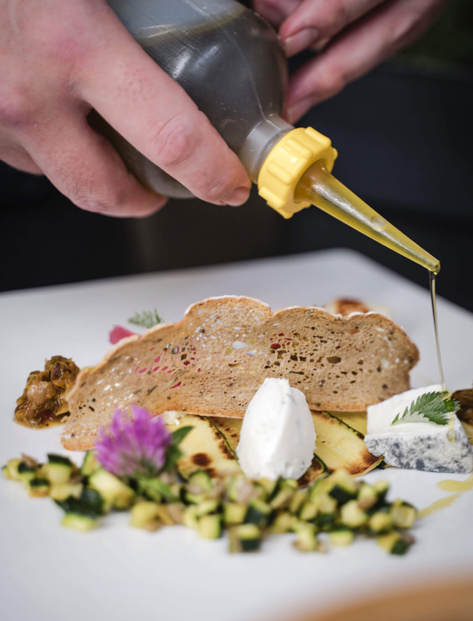 A hand pours oil onto a gourmet dish with toast, cheese, grilled zucchini, and edible flowers on a white plate. - Hotel Moosmair