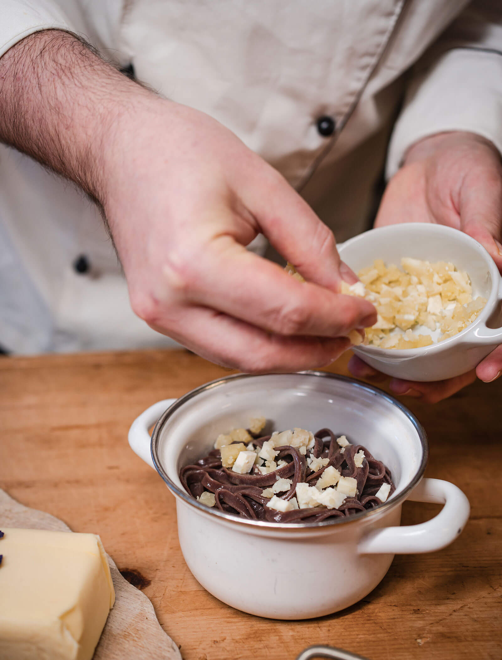 A chef sprinkles cheese over a bowl of pasta on a wooden counter. - Hotel Moosmair