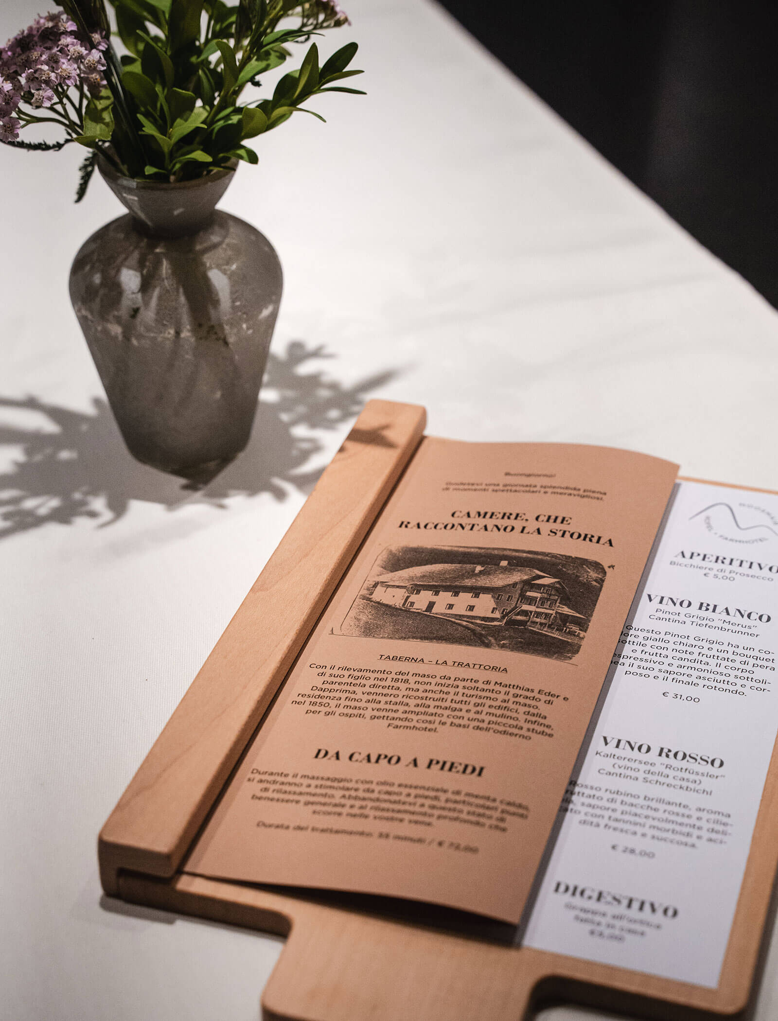 A menu on a wooden board next to a small vase with flowers on a white tablecloth. - Hotel Moosmair