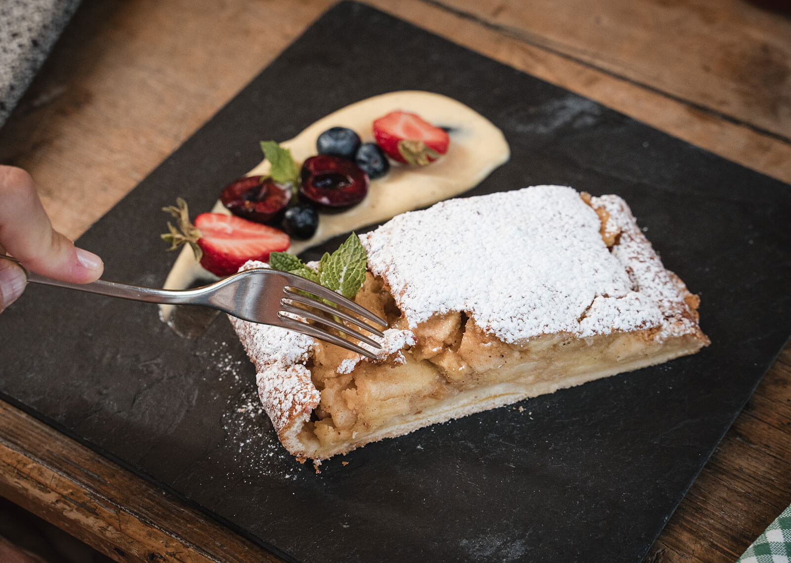 A fork cutting into apple strudel dusted with powdered sugar, served with cream and berries on a slate plate. - Hotel Moosmair
