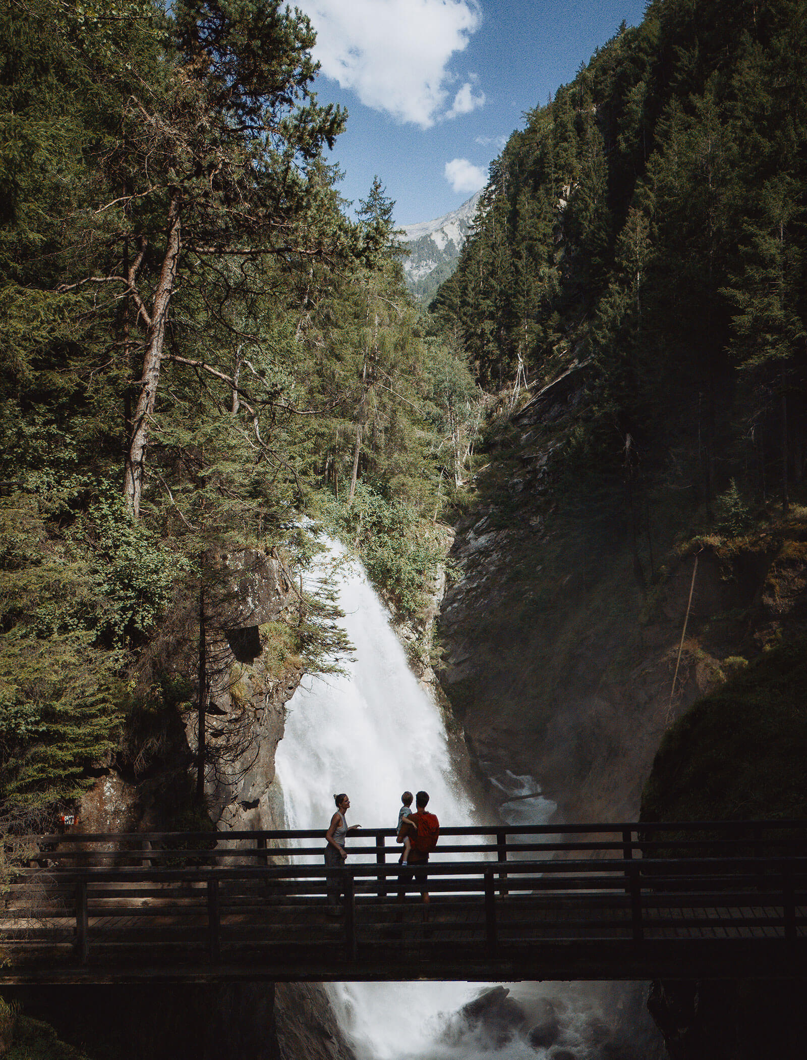 Three people stand on a wooden bridge overlooking a waterfall surrounded by trees and mountains. - Hotel Moosmair