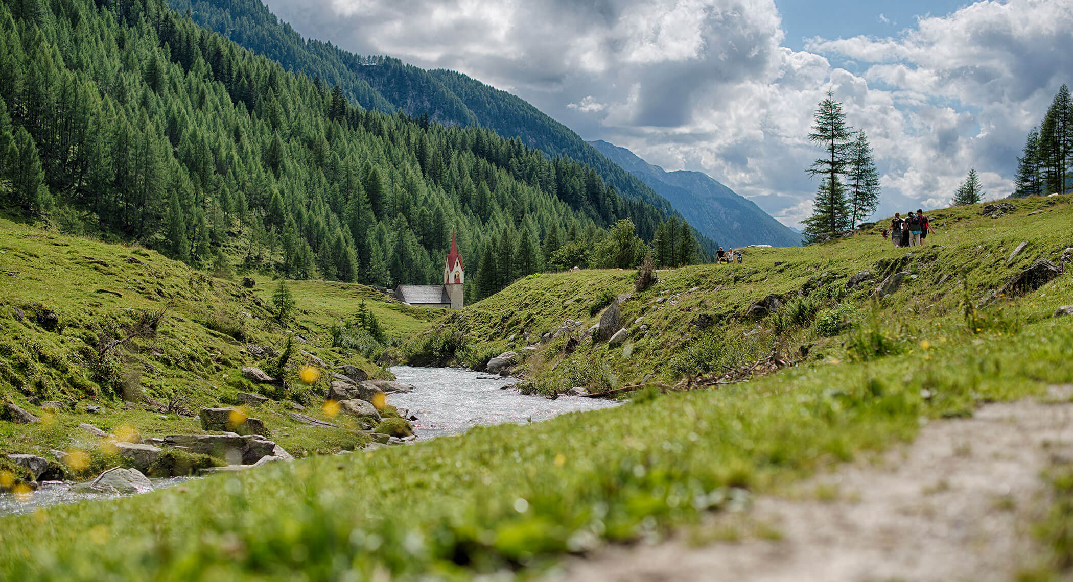 A small red-roofed chapel by a stream in a green valley with forested mountains and a cloudy sky. - Hotel Moosmair