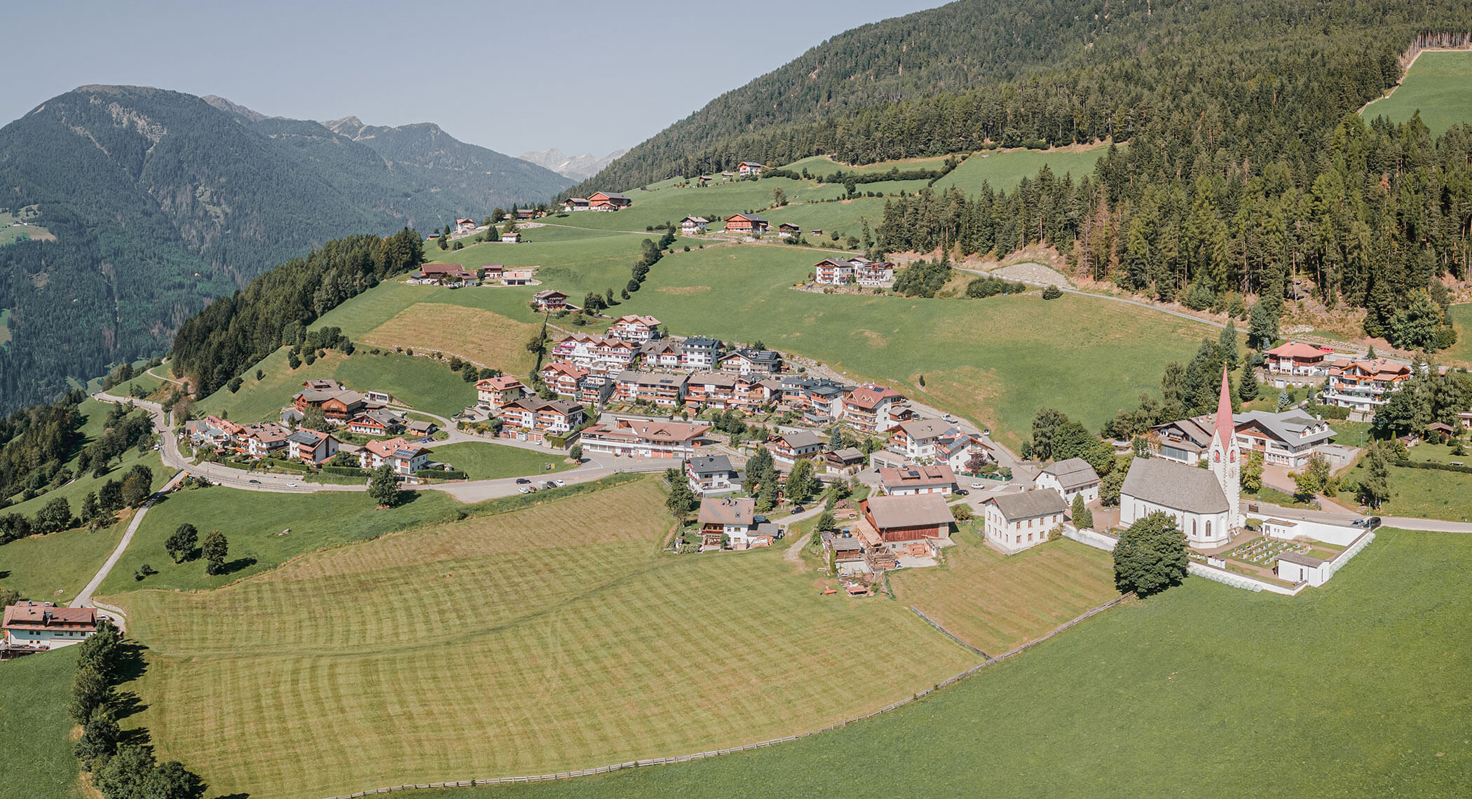 Aerial view of a mountain village with scattered houses, green fields, and forested hills. - Hotel Moosmair