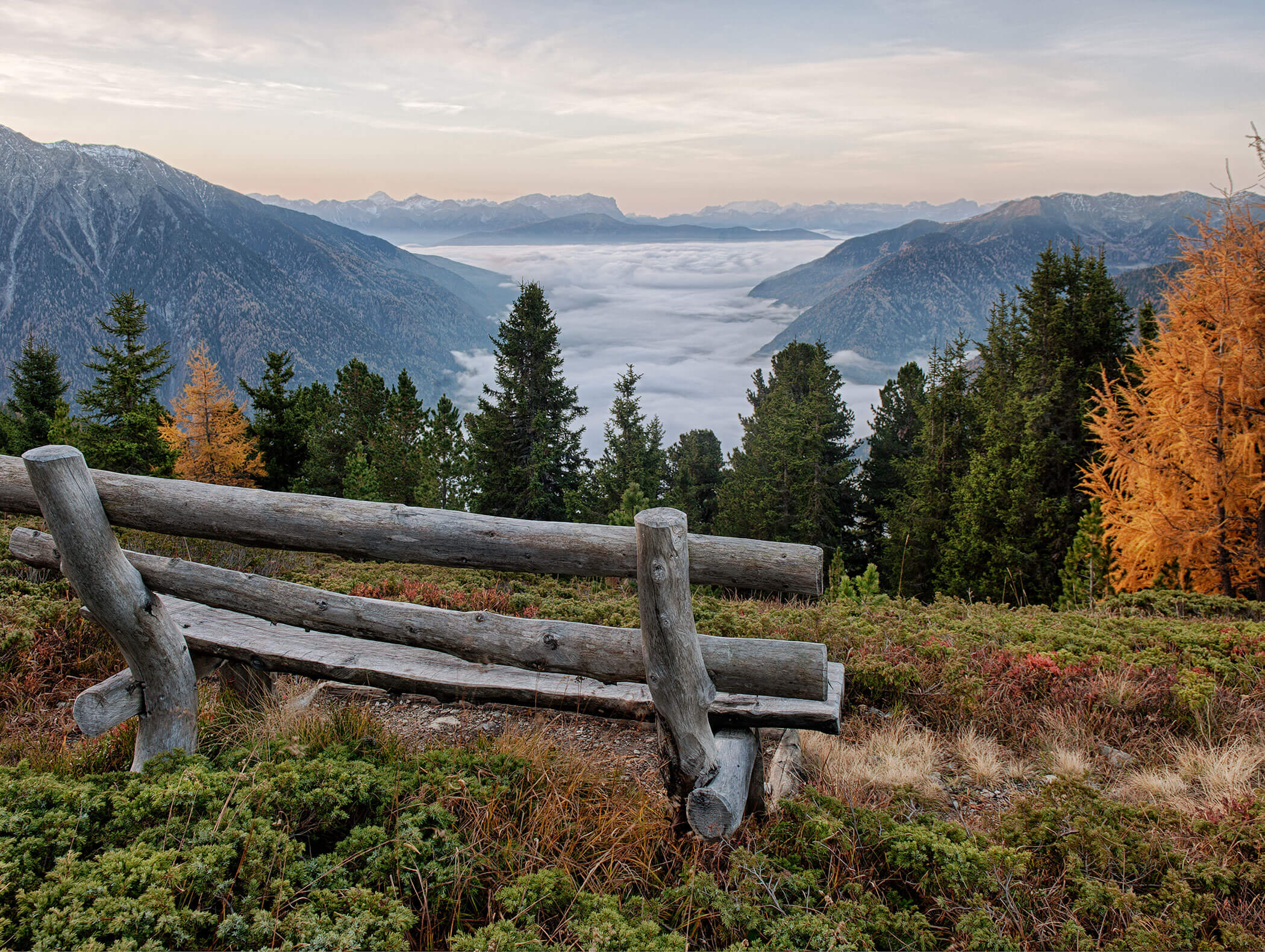 A rustic wooden bench overlooks mist-filled valleys, mountains, and autumn trees under a soft sky. - Hotel Moosmair
