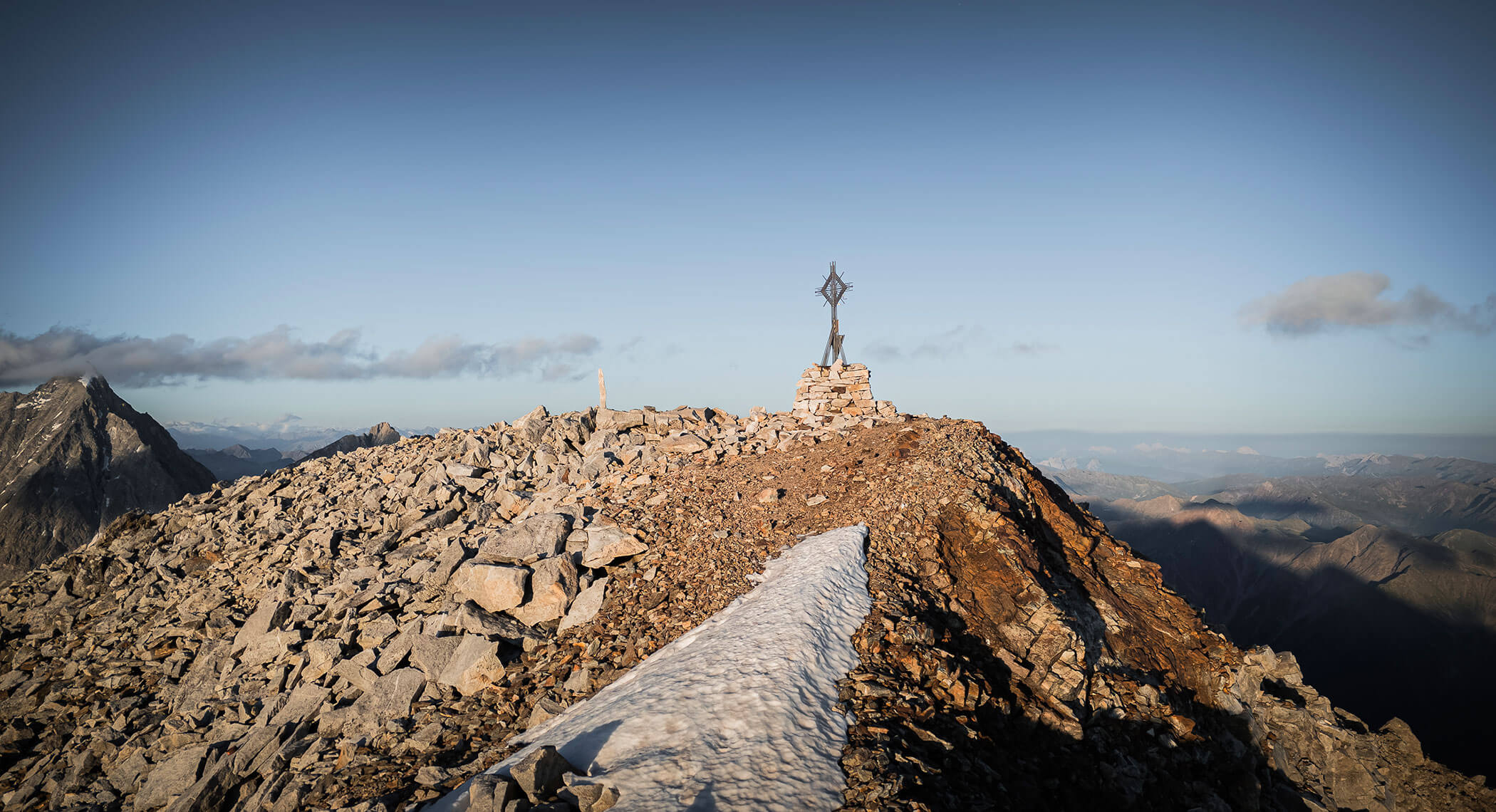 Rocky mountain peak with a statue on top, patches of snow, and distant mountains under a clear sky. - Hotel Moosmair