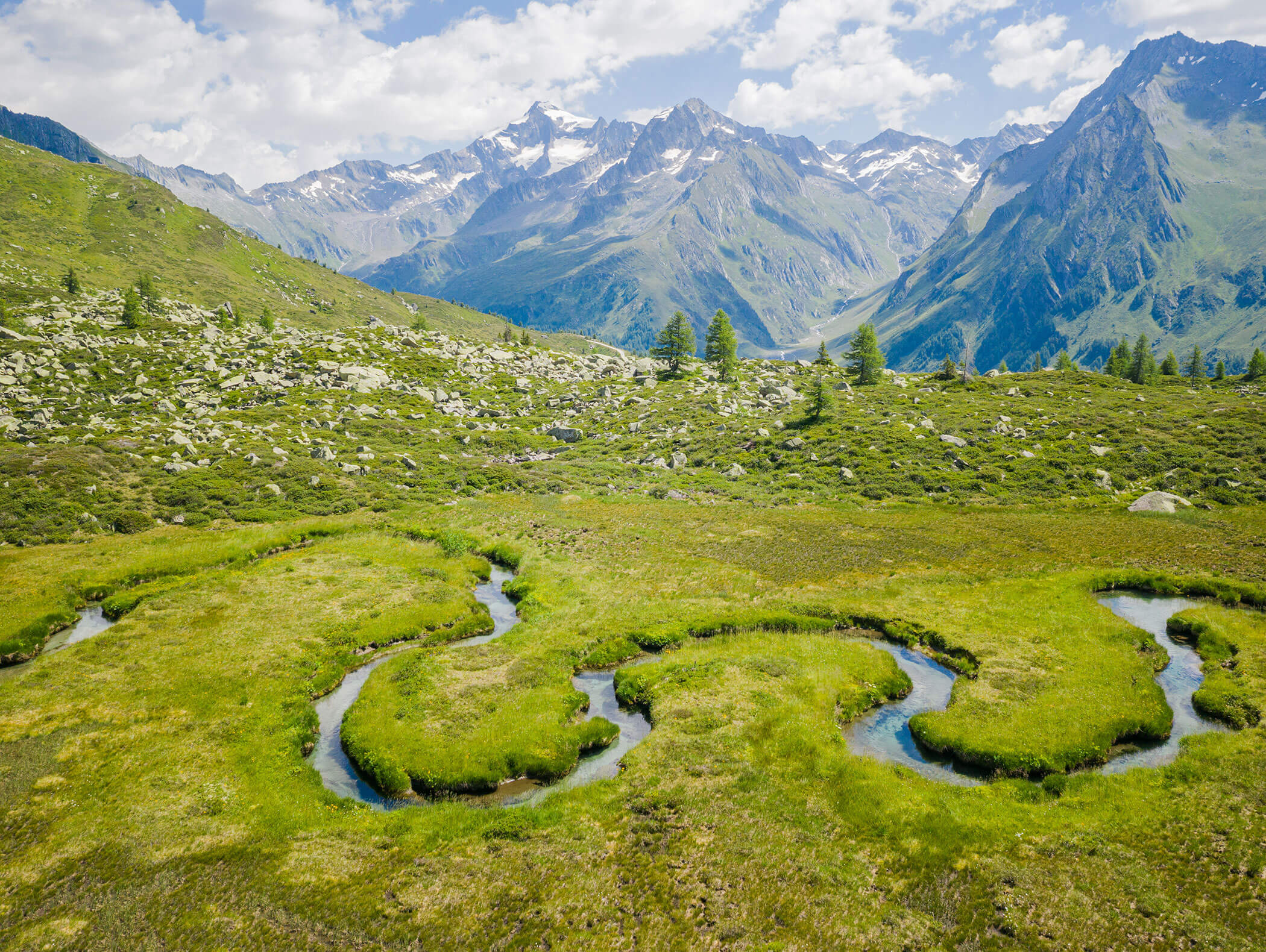 Winding stream through green meadow with mountains and partly cloudy sky in the background. - Hotel Moosmair