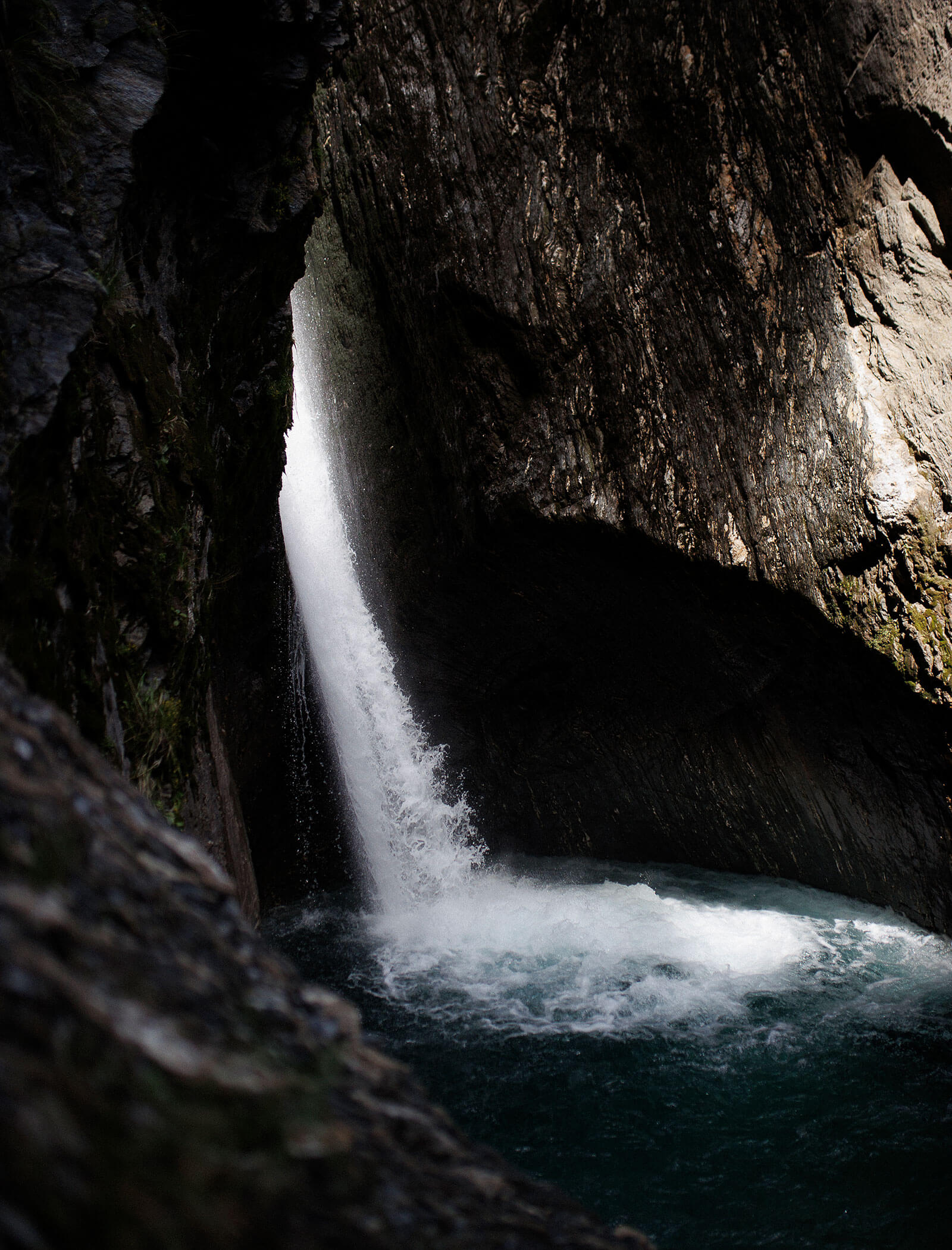 A narrow waterfall flows between dark rock cliffs into a turbulent blue pool below. - Hotel Moosmair