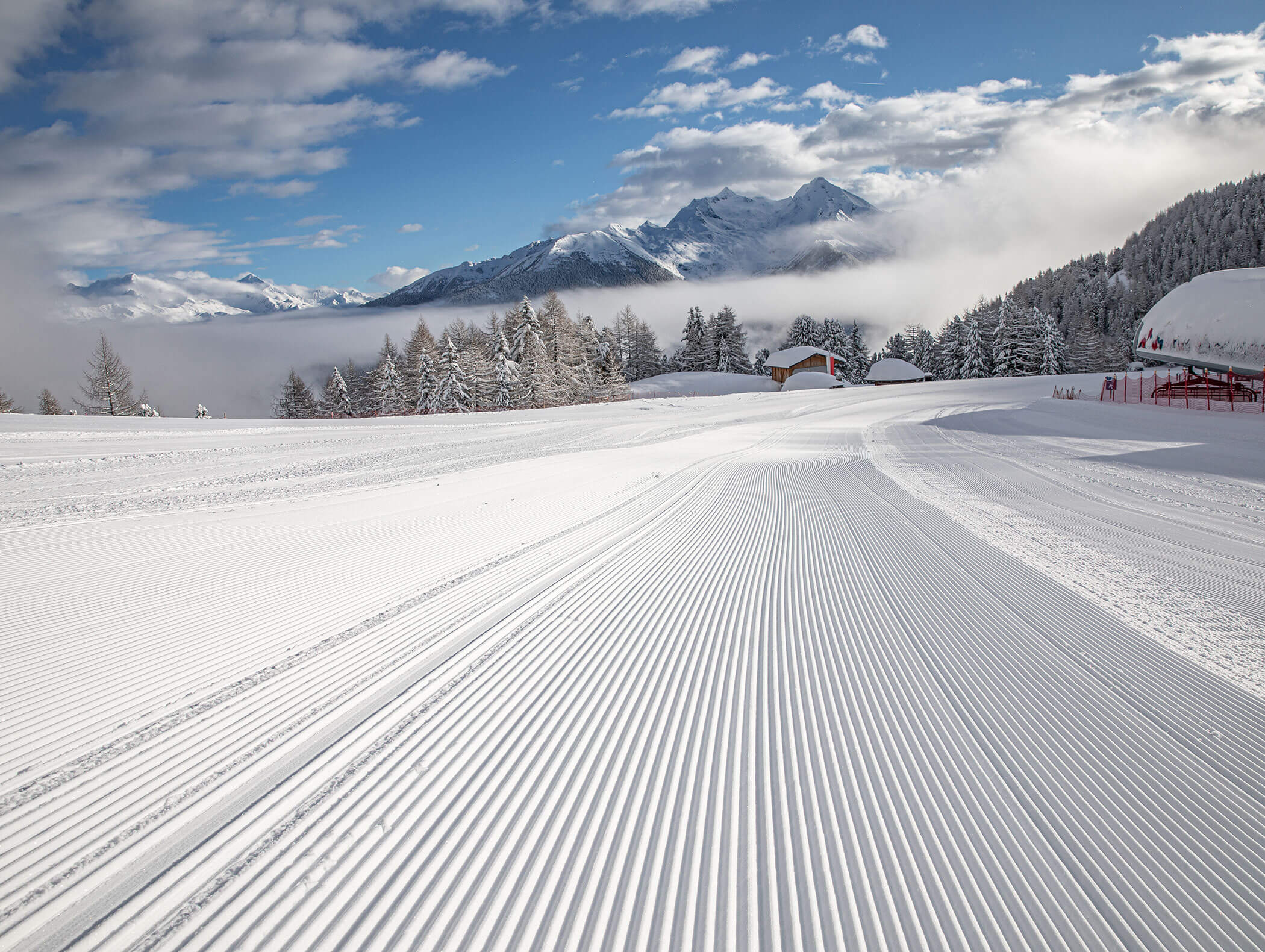 Neatly groomed ski slope with snowy trees, mountains, and clouds under a blue sky in the background. - Hotel Moosmair
