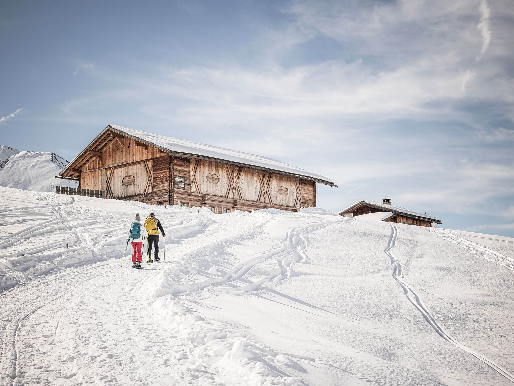 Two people walk in deep snow toward wooden cabins on a sunny, snowy mountain slope under a blue sky. - Hotel Moosmair