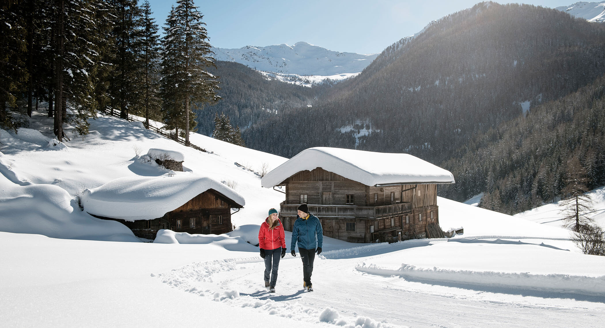 A couple walks on a snowy path near wooden cabins in a mountain landscape on a sunny winter day. - Hotel Moosmair