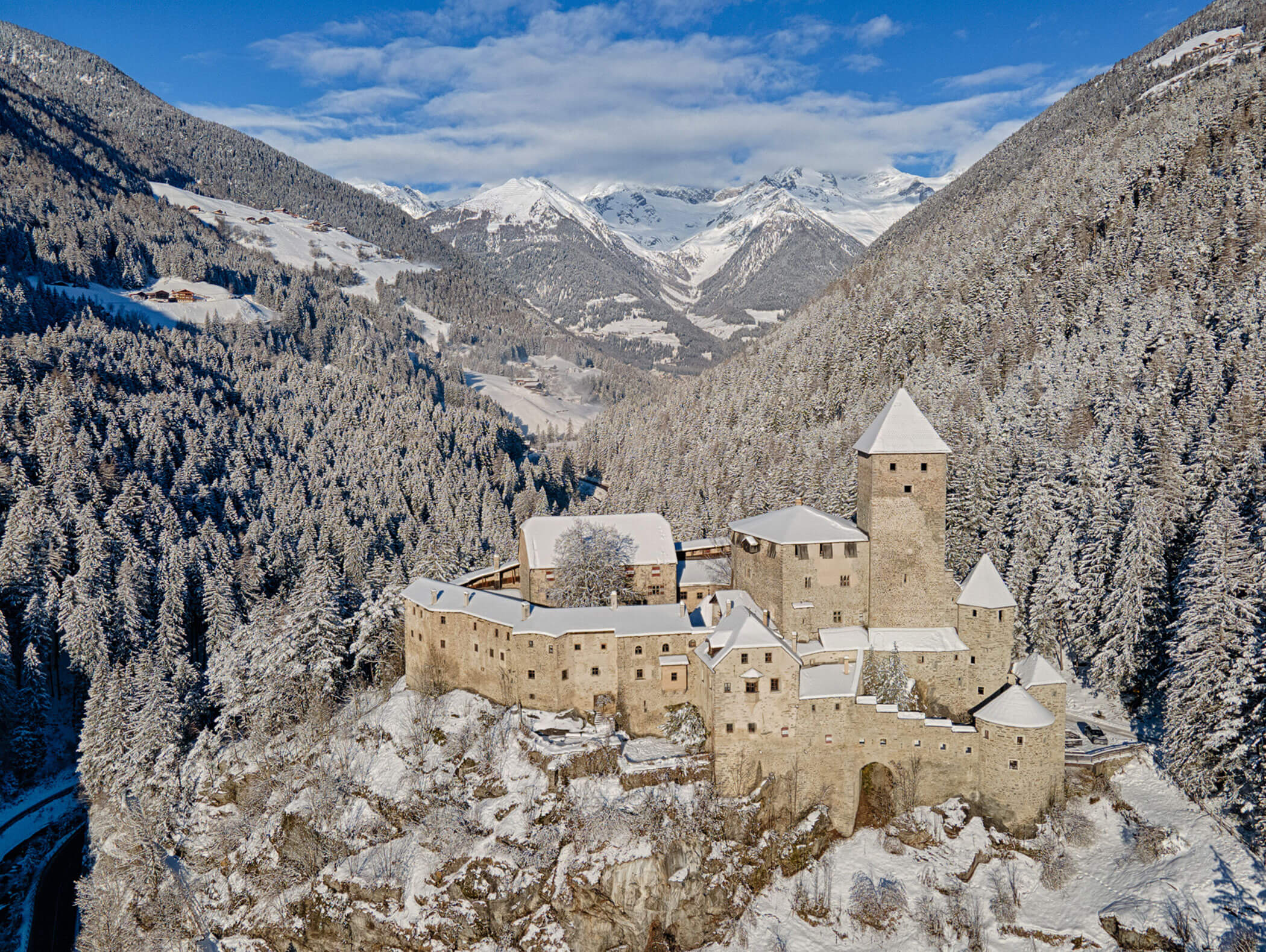 Medieval stone castle on a snowy hill, surrounded by forested mountains under a blue sky. - Hotel Moosmair