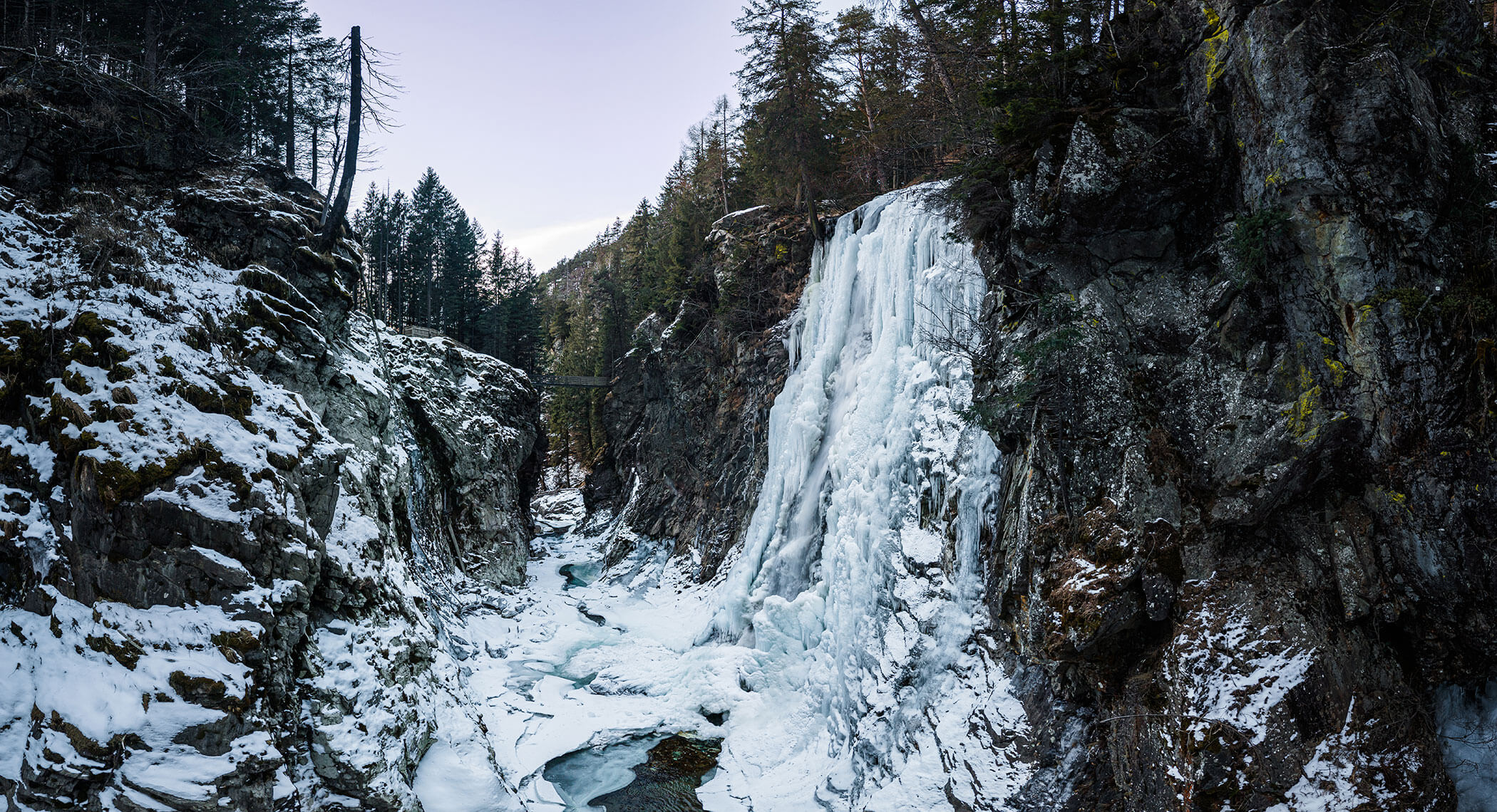 A large frozen waterfall cascades down rocky cliffs surrounded by snow and evergreen trees. - Hotel Moosmair