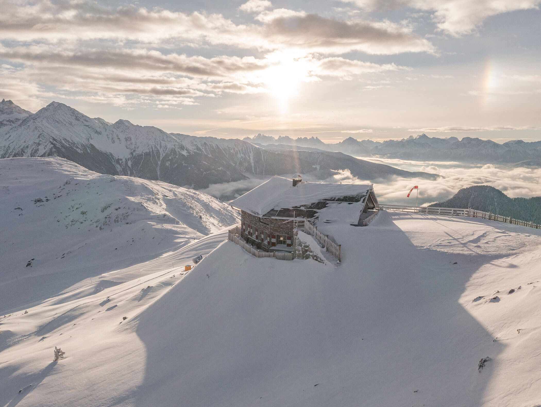 Snow-covered mountain hut with surrounding peaks and sunrise, overlooking a misty valley below. - Hotel Moosmair