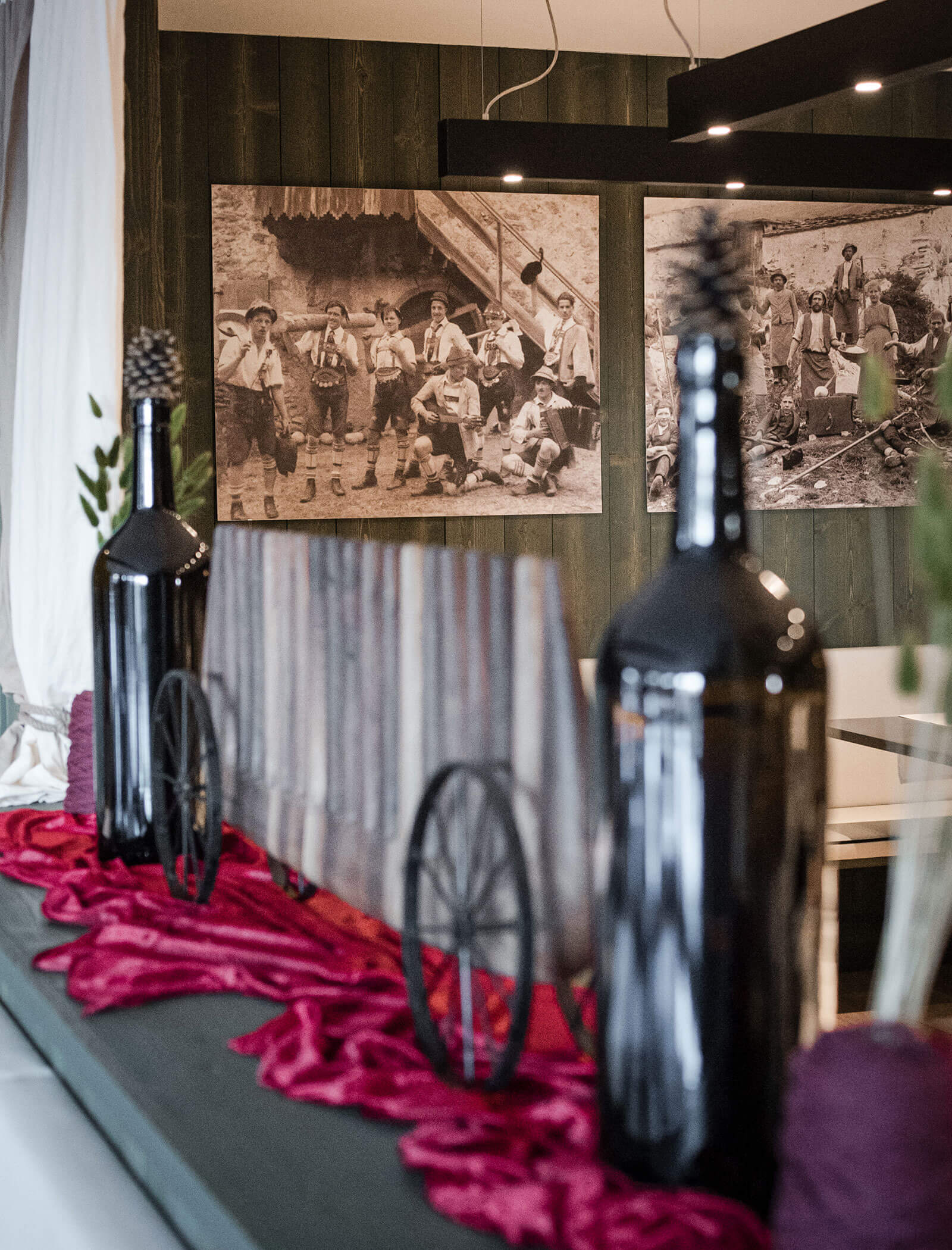 Due bottiglie scure e un piccolo carrello su un tavolo con stoffa rossa, foto d'epoca sulla parete di legno alle spalle. - Hotel Moosmair
