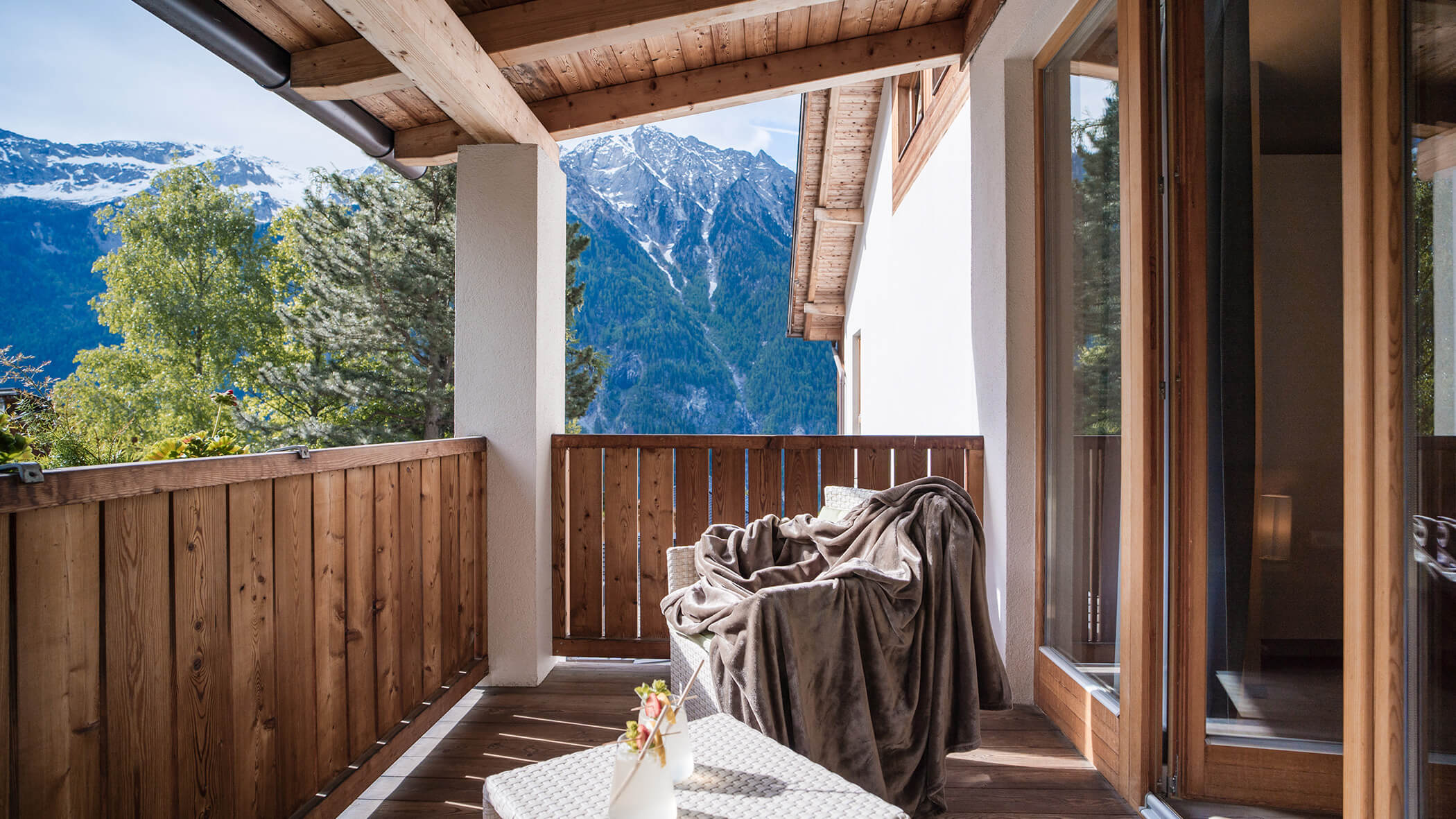 Un accogliente balcone in legno con una sedia coperta e vista sulle montagne sotto un cielo parzialmente nuvoloso. - Hotel Moosmair