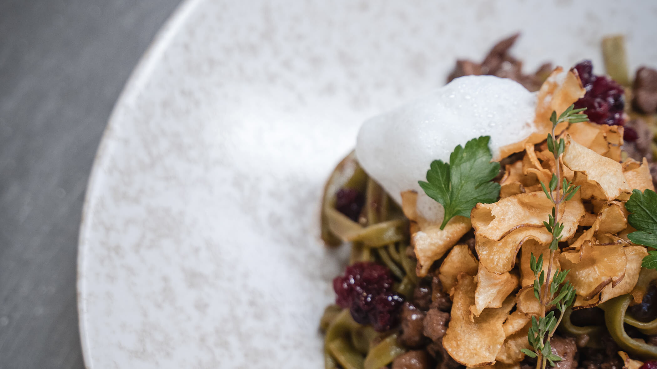 Gourmet pasta dish topped with foam, herbs, crispy chips, and a rich sauce on a speckled plate. - Hotel Moosmair