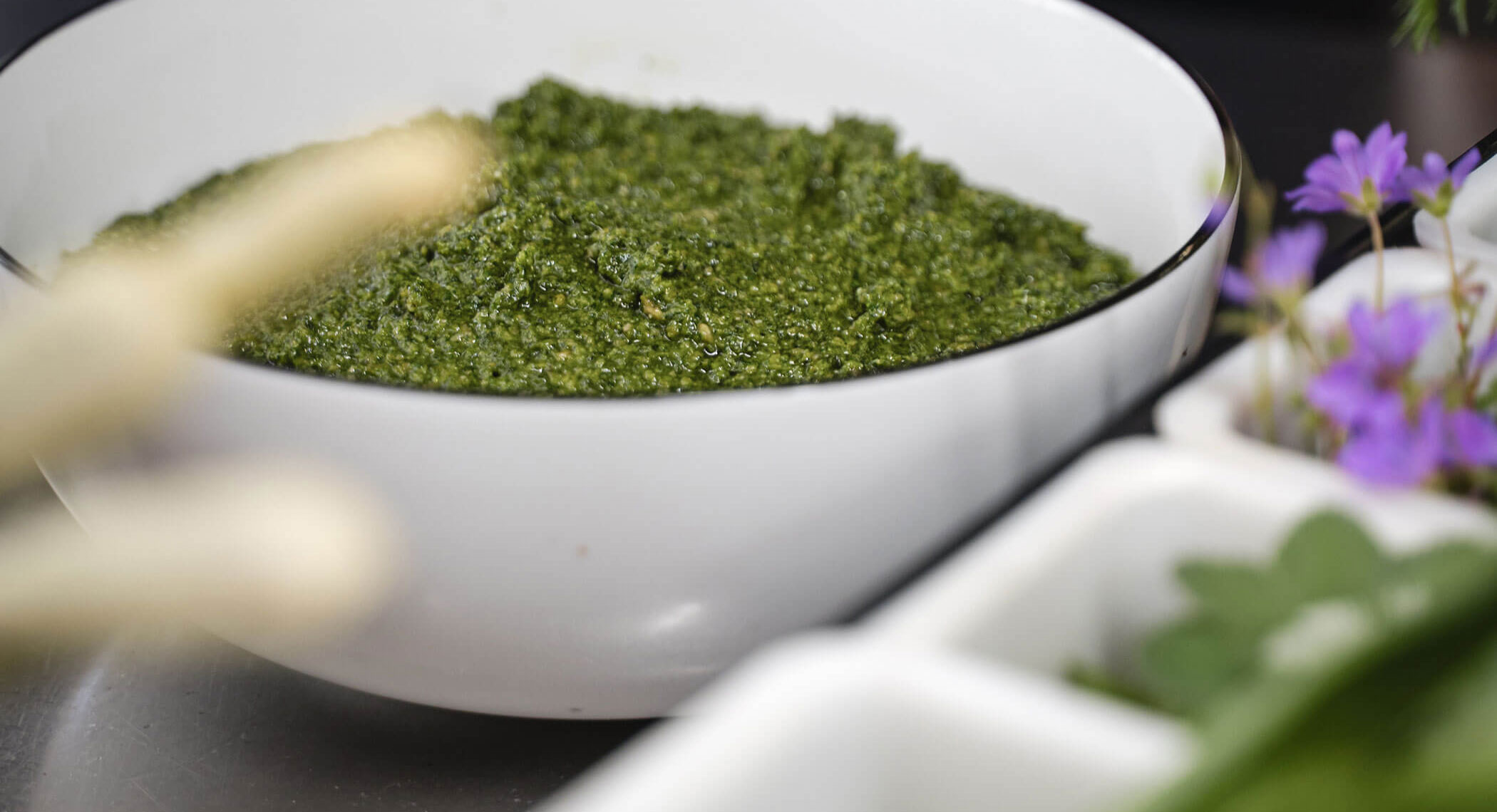 Una ciotola bianca piena di pesto verde, circondata da erbe e piccoli fiori viola. - Hotel Moosmair