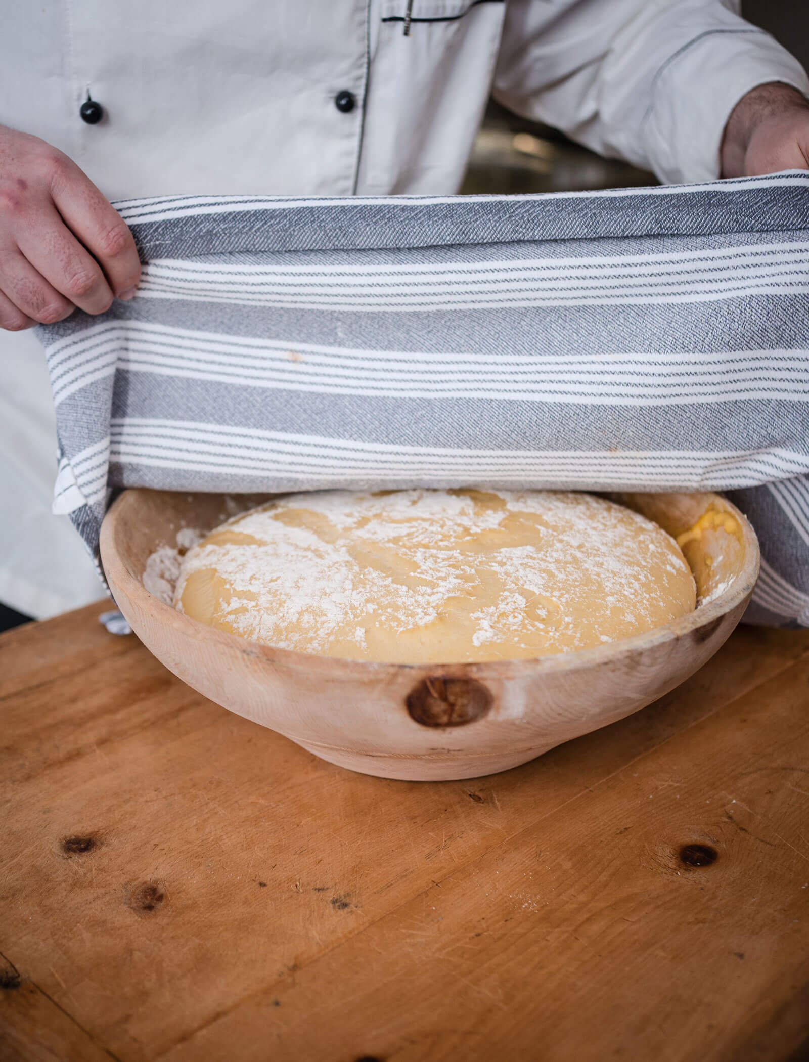 Una persona copre una ciotola di pasta in lievitazione con un panno a righe su un tavolo di legno. - Hotel Moosmair