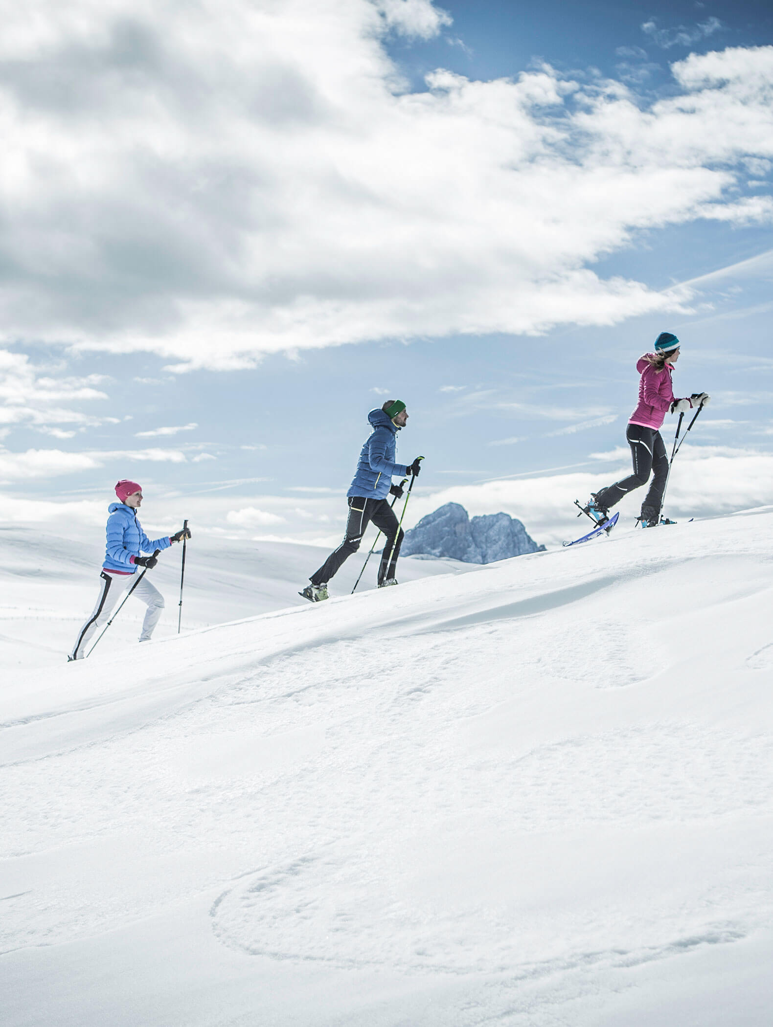 Drei Personen in Winterkleidung gehen mit Schneeschuhen auf einen verschneiten Hügel unter einem teilweise bewölkten Himmel mit Bergen im Hintergrund. - Hotel Moosmair