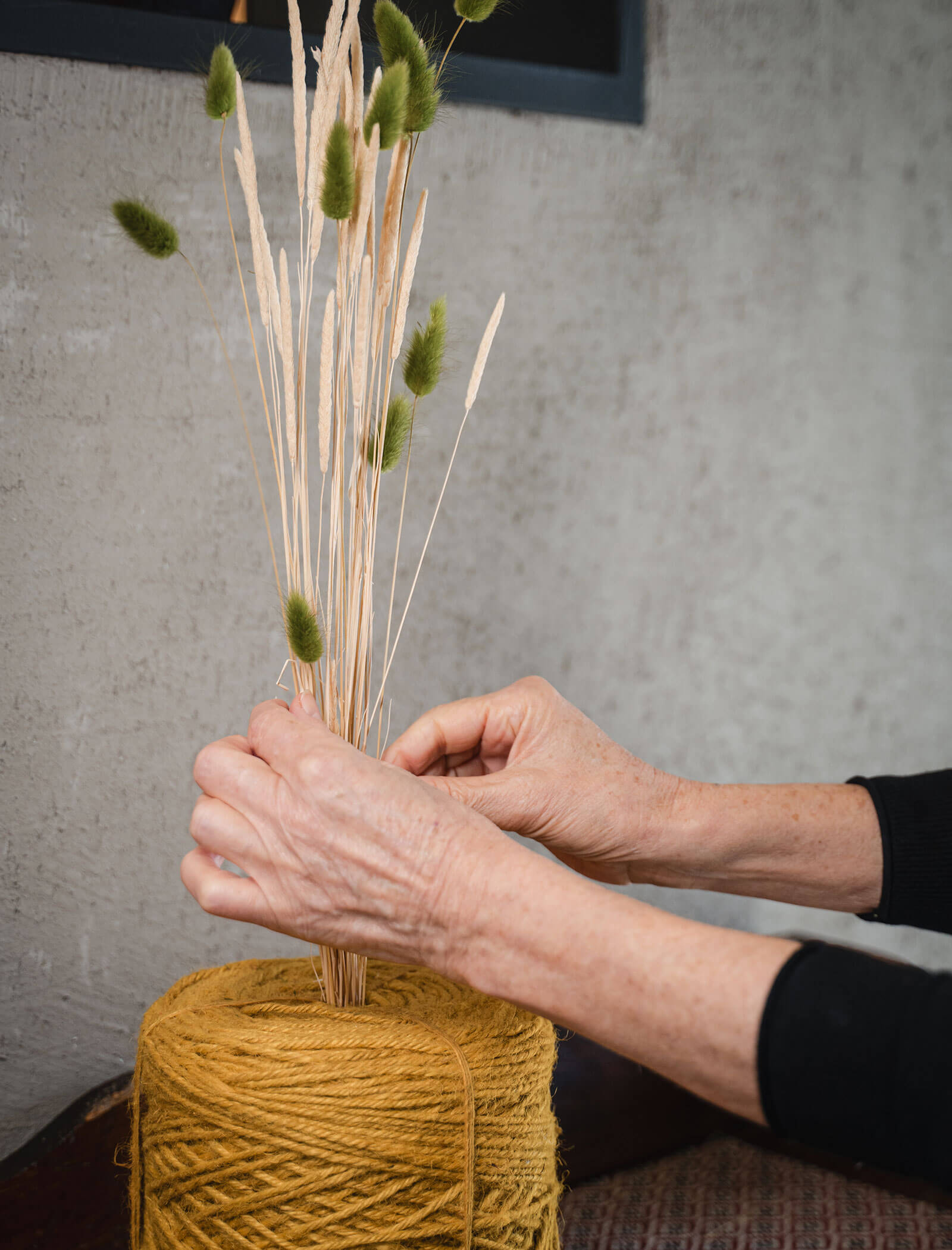 Hände, die getrocknete Gräser in einer Vase arrangieren, die mit dickem gelben Garn umwickelt ist, vor einer grauen Wand. - Hotel Moosmair